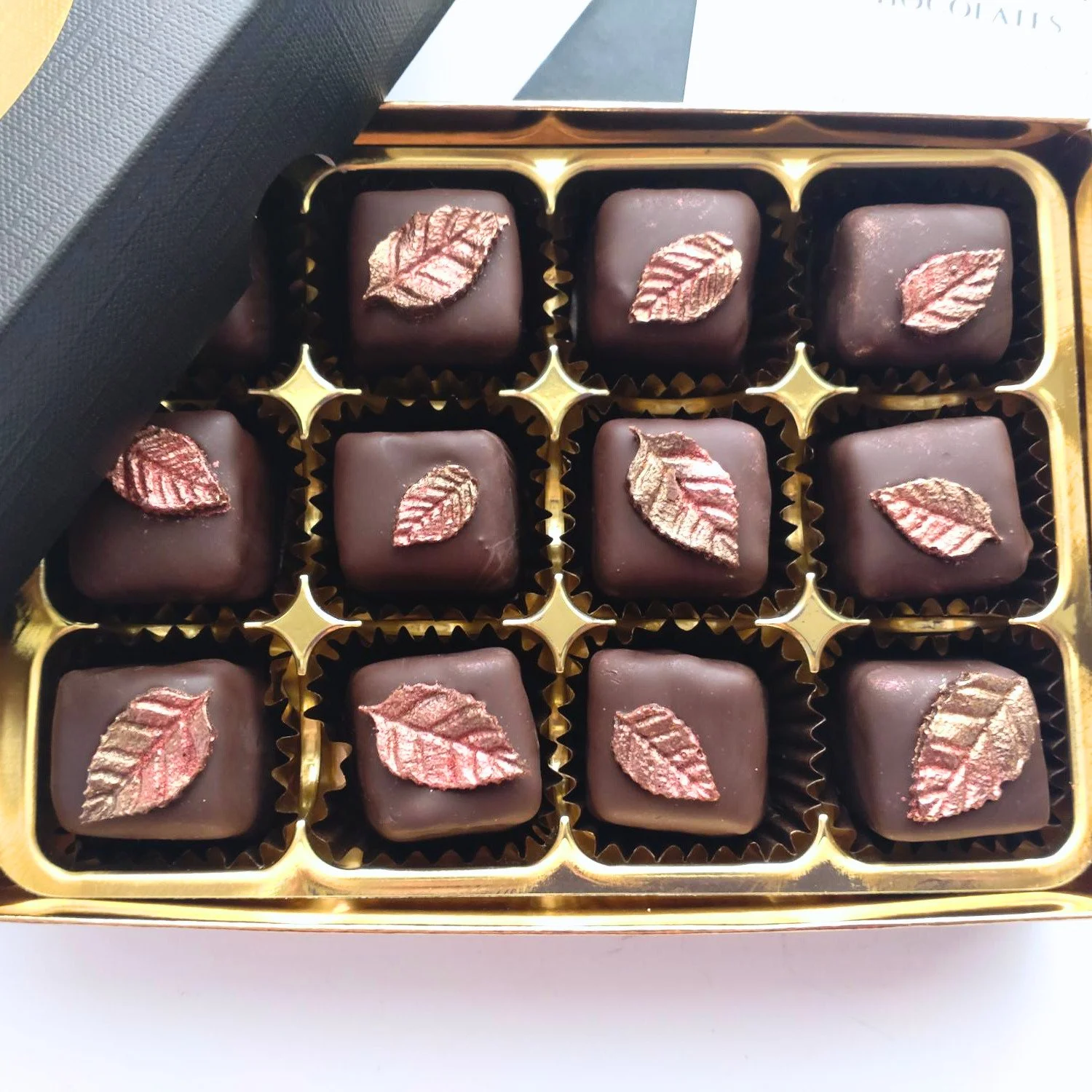 Close-up of twelve handmade dark chocolates in a gold gift tray, each topped with a bronze and copper-coloured chocolate leaf.