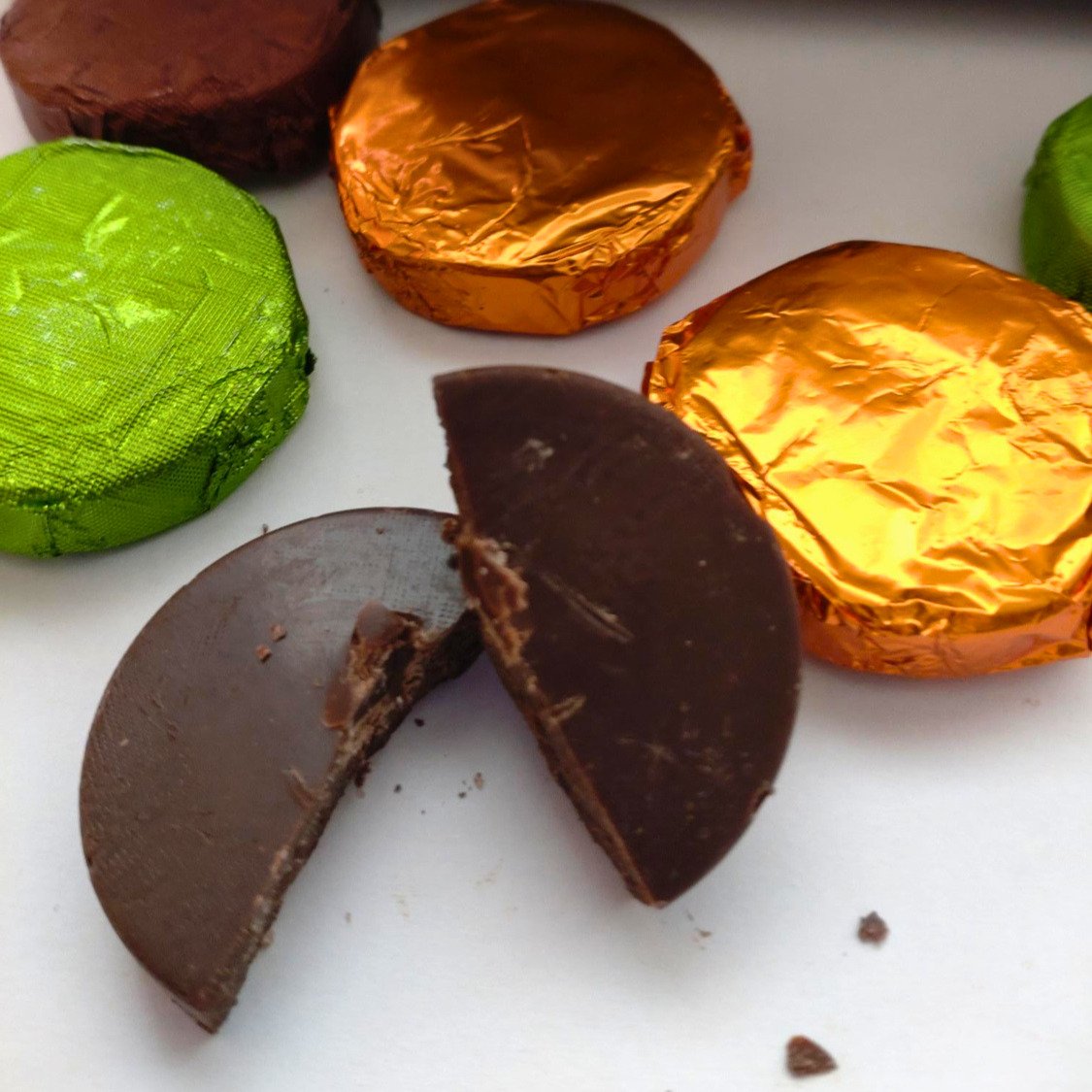 Chocolate discs wrapped in orange, brown and green foil on a white background. One dark chocolate disc is unwrapped and broken in half.