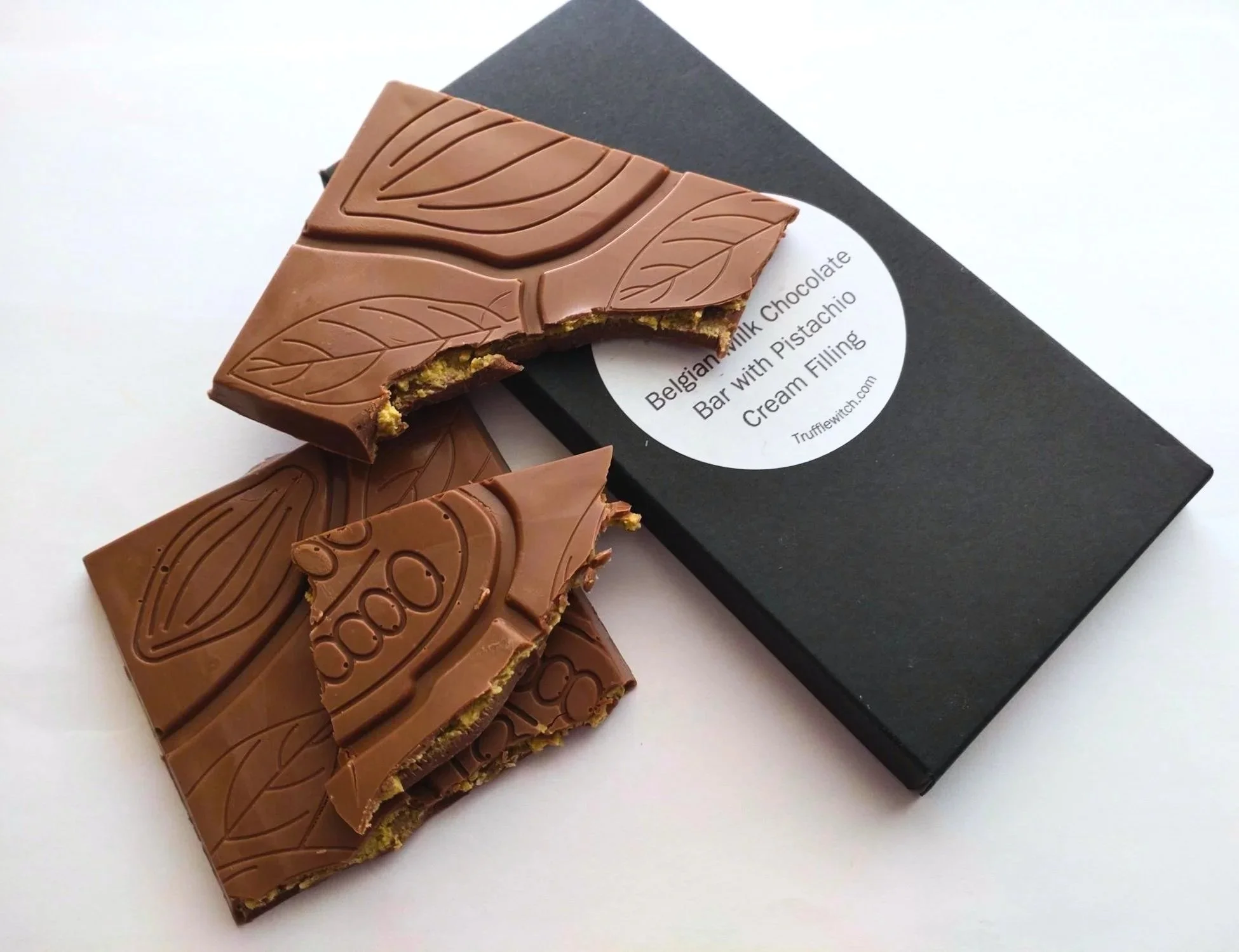 Milk chocolate bar with pistachio cream filling broken into 3 pieces laid on top of a black chocolate bar carton with a white label