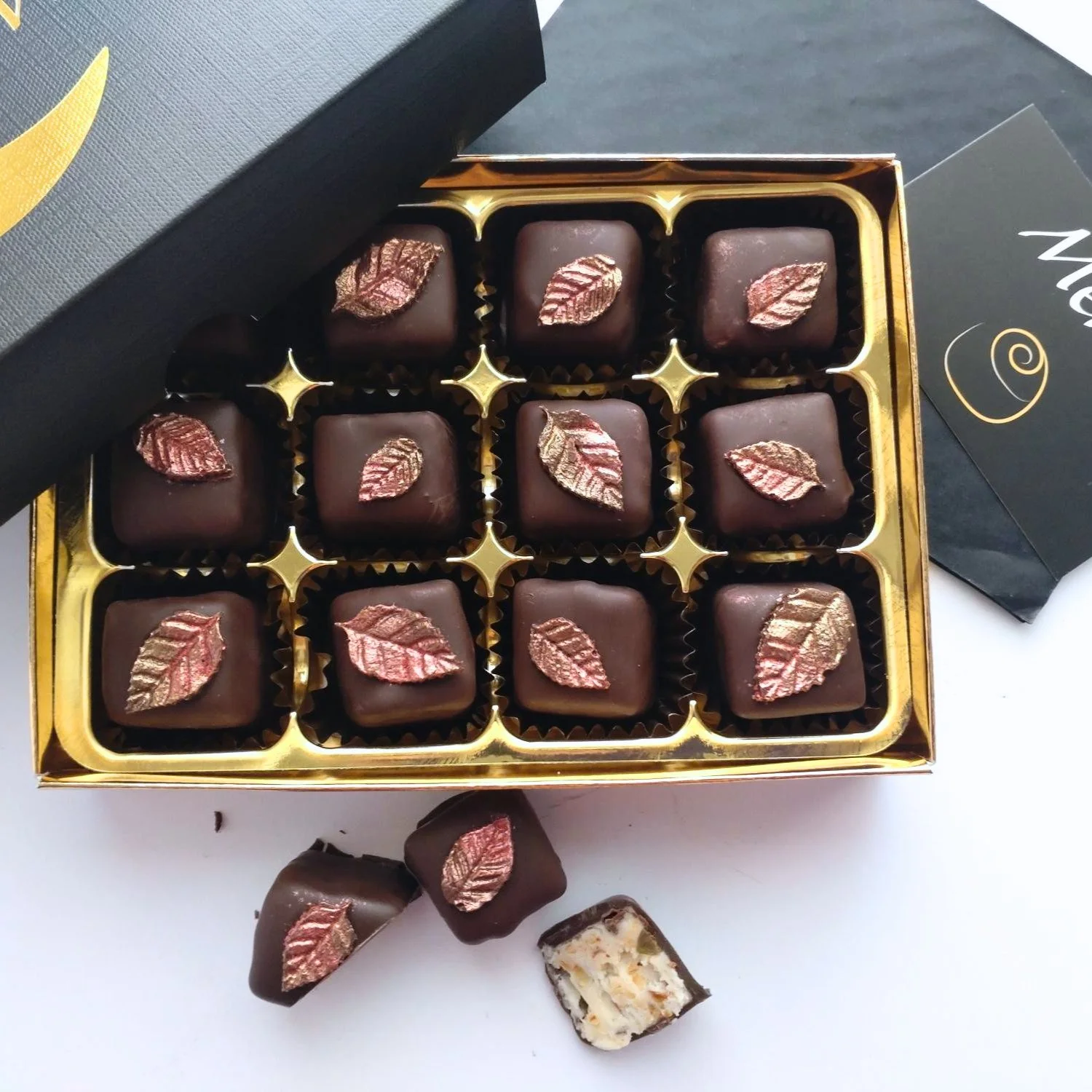 Open Truffle Witch gift box containing twelve Taste of Autumn chocolates decorated with metallic bronze leaves, with cut chocolates showing the nutty white chocolate filling in front.