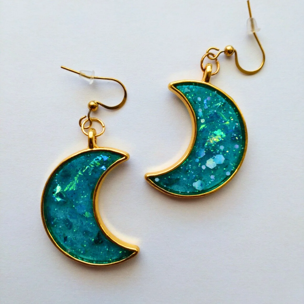 Gold coloured crescent moon earrings in glittery turquoise resin on white background