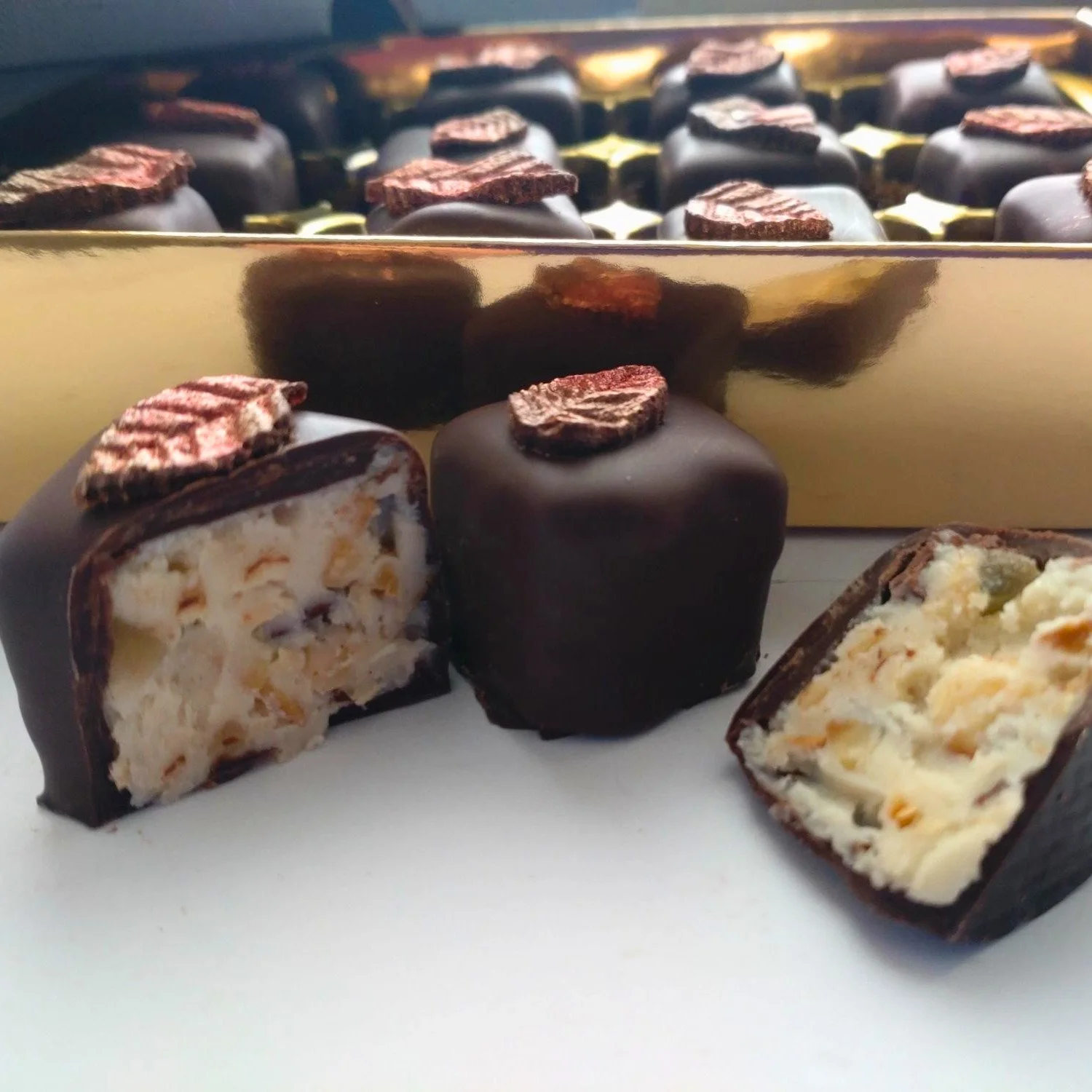 Low-angle view of handmade dark chocolates decorated with metallic autumn leaves, with cut pieces displaying their textured white chocolate centres in front of a gold gift tray.
