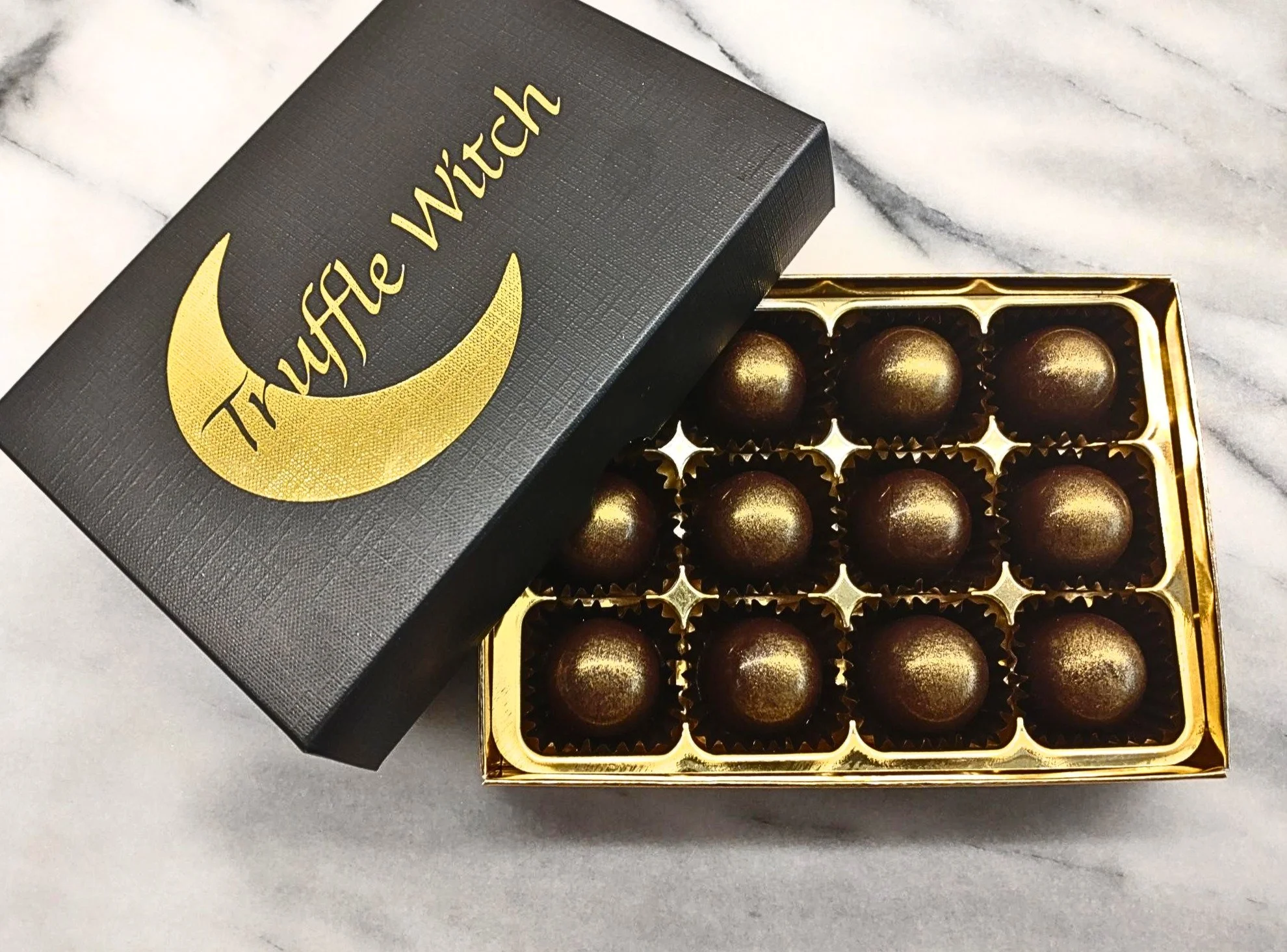 Open box of caramels on a marble backgound. Box has a black lid with Truffle Witch logo in gold foil. The caramels are in black pettit four cases and dusted with gold lustre.