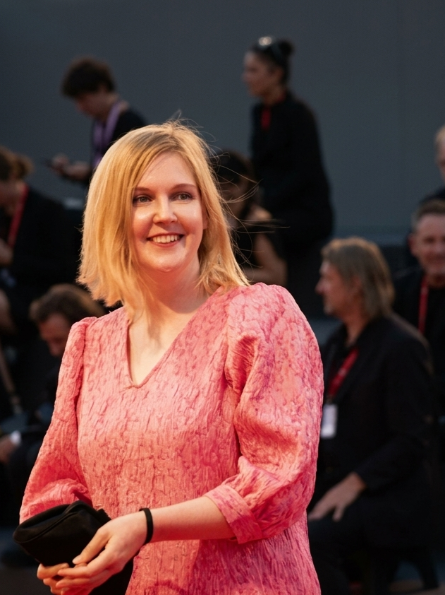 Filmmaker Hannaleena Hauru smiling on the red carpet at the Venice Biennale, wearing a textured pink dress and holding a black clutch bag.