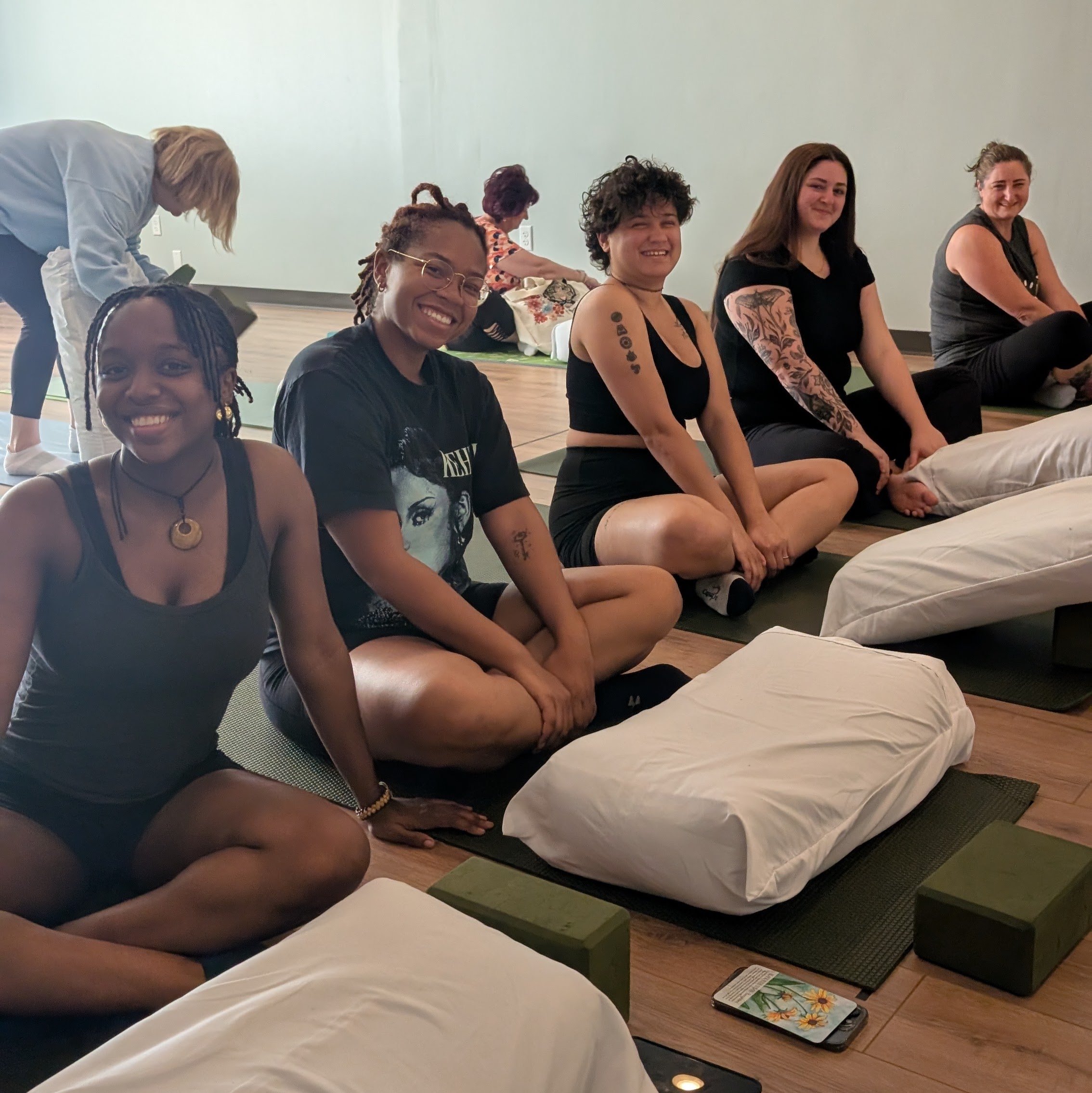 Smiling yoga students sitting on bolsters before class at Emerald Yoga Studio in Pembroke, MA, near Kingston