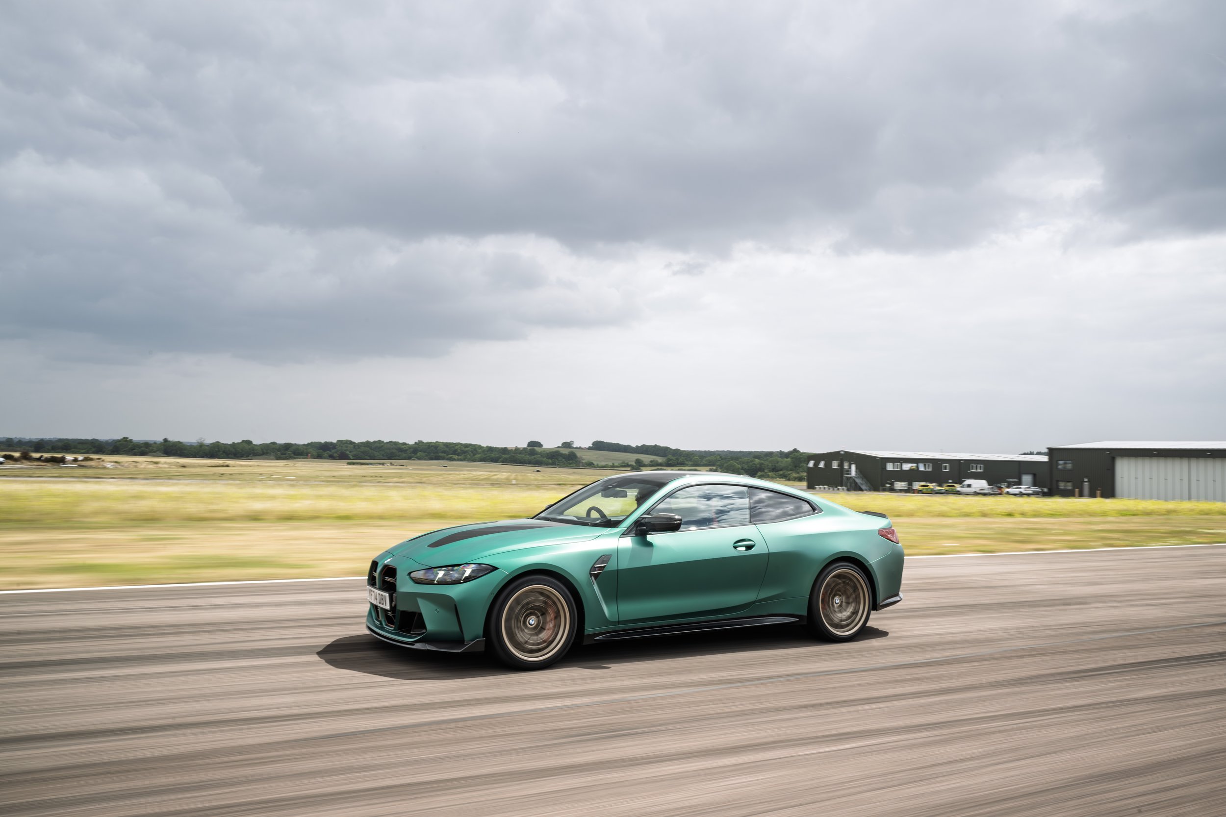 A green sports car driving on an open road with a cloudy sky above and some industrial buildings in the background.