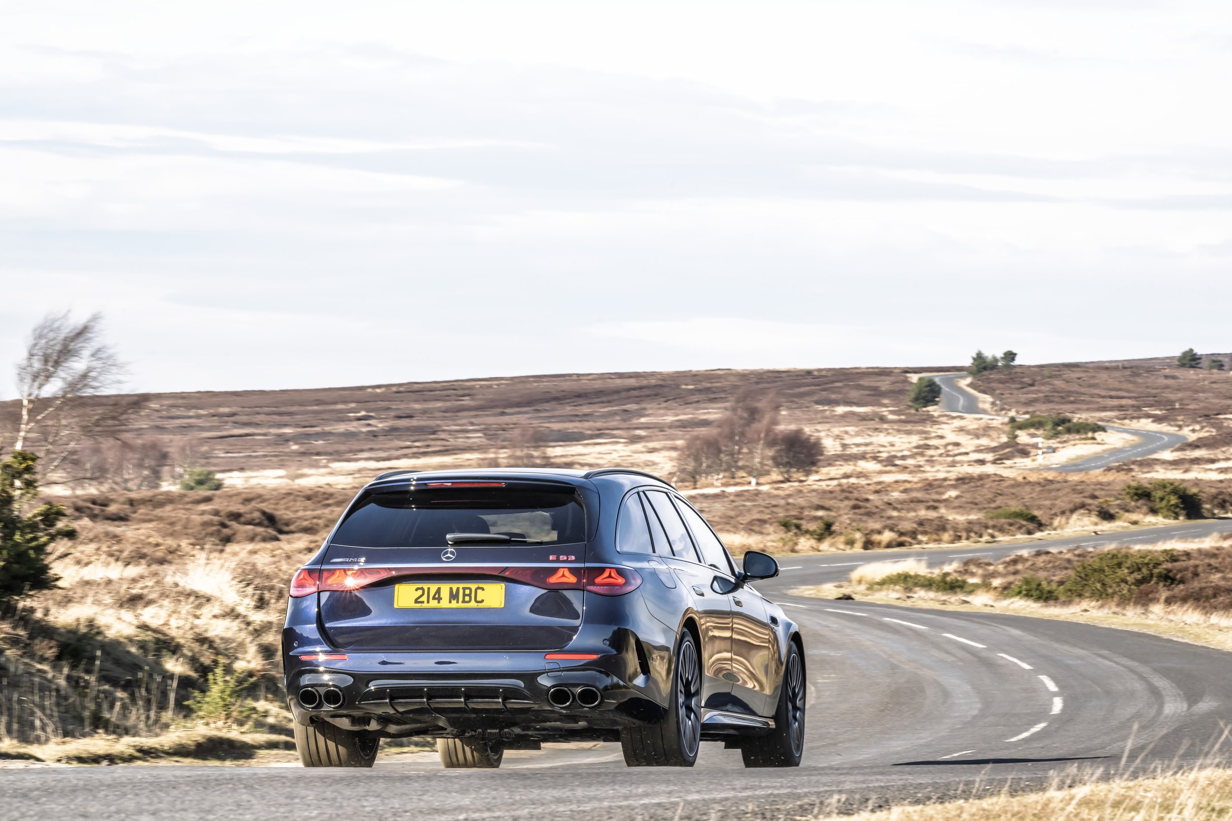 A black Mercedes-Benz E 53 AMG station wagon driving on a winding country road through a rural landscape with dry grass and hills in the background.