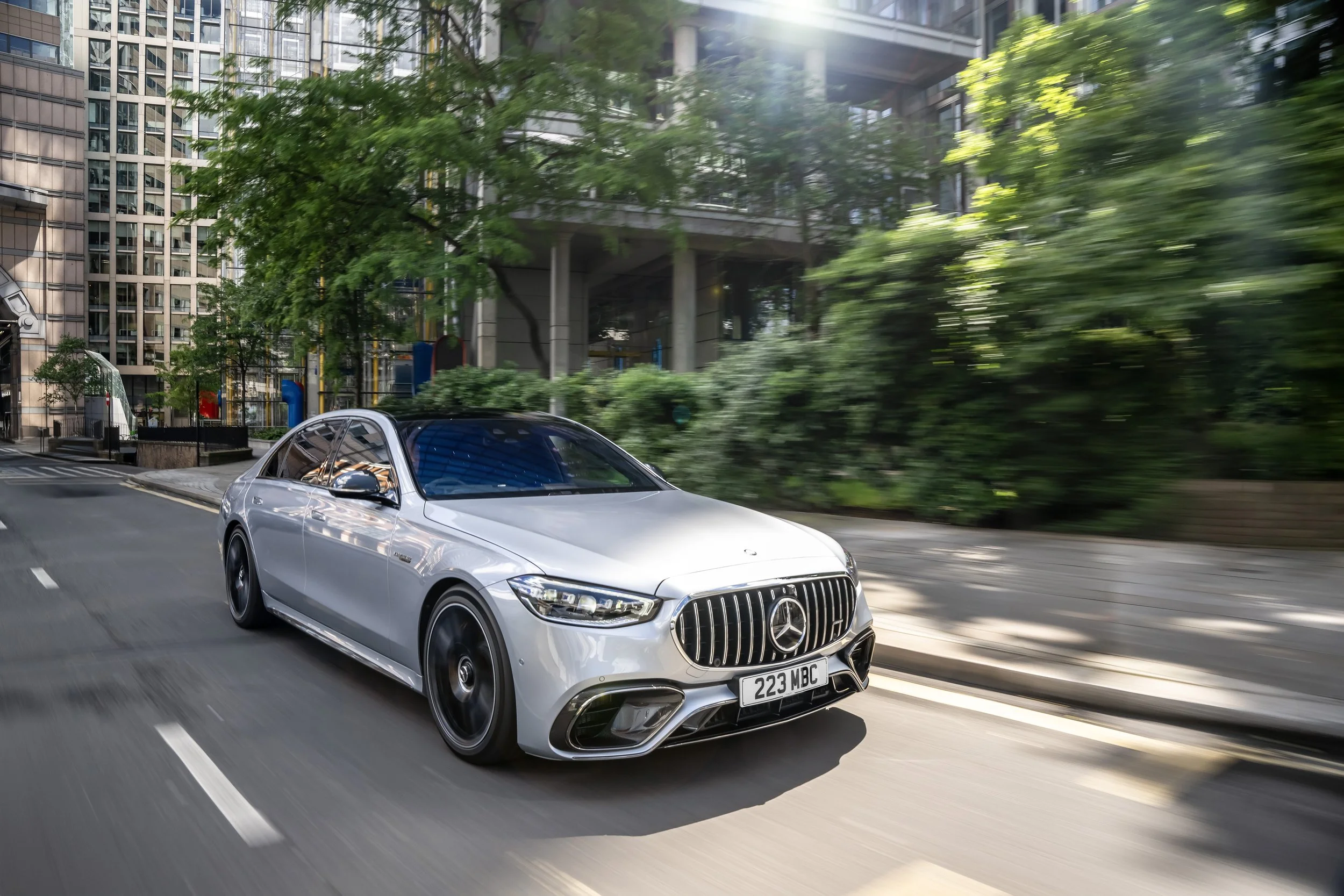 Silver Mercedes-Benz car driving on city street surrounded by green trees and tall buildings.