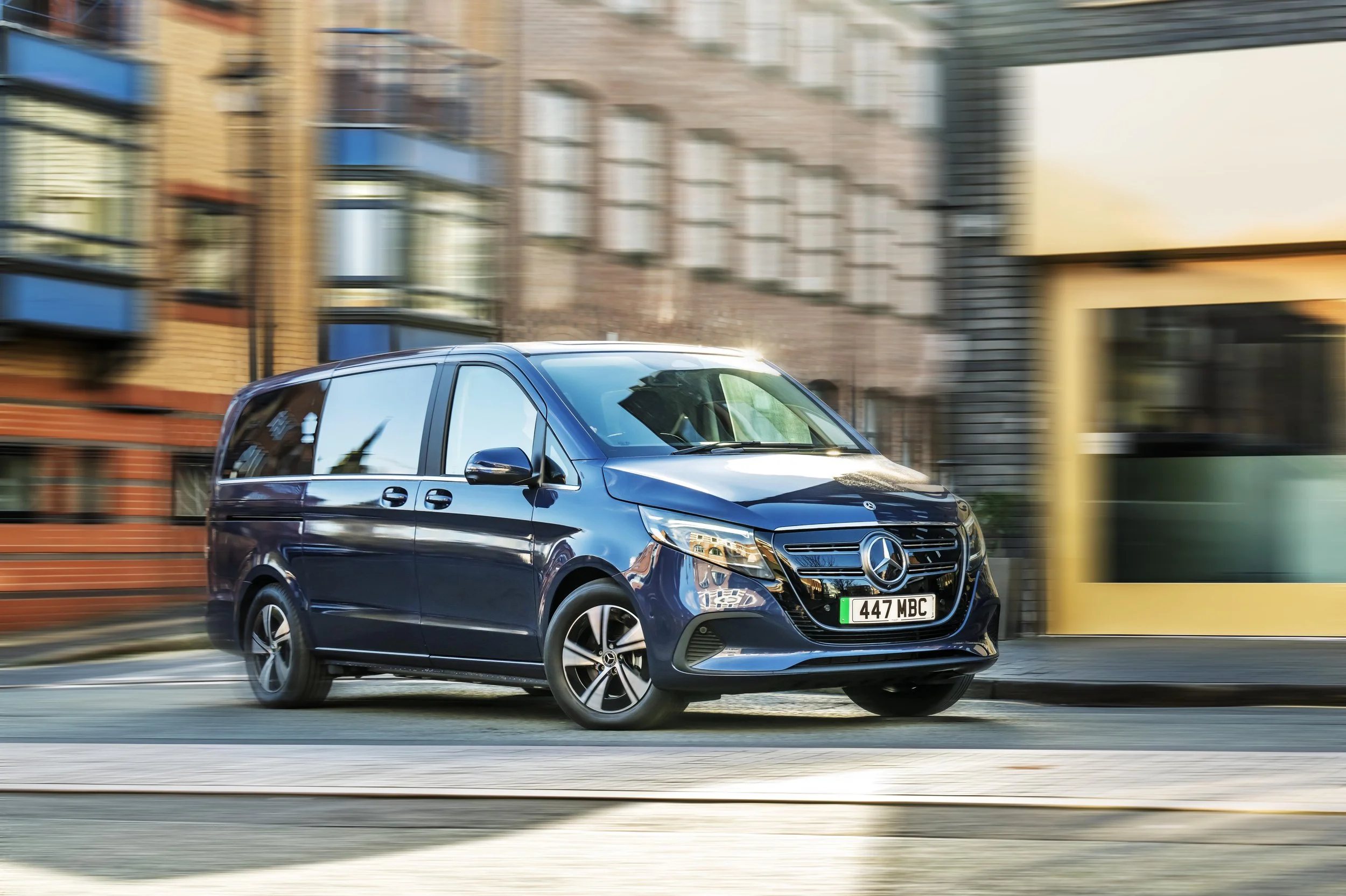 A blue Mercedes-Benz van driving on a city street with blurred buildings and storefronts in the background.