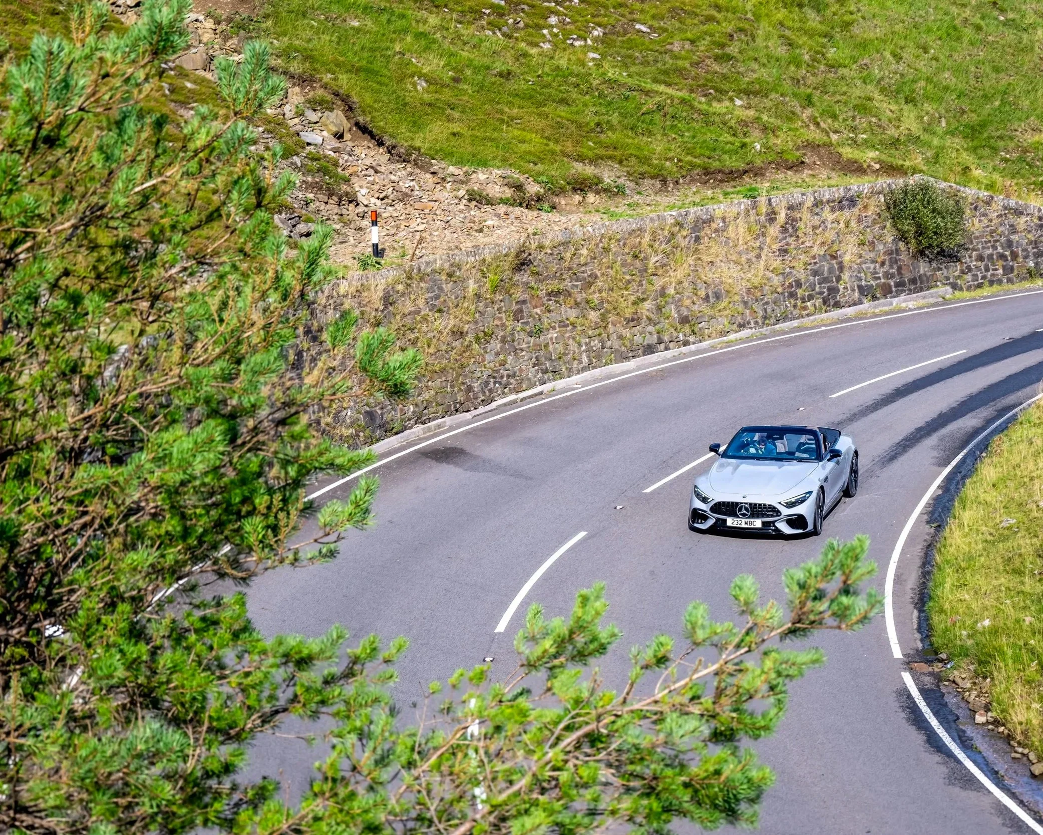 A silver convertible Mercedes-Benz driving on a winding mountain road with green grassy hills and trees around.
