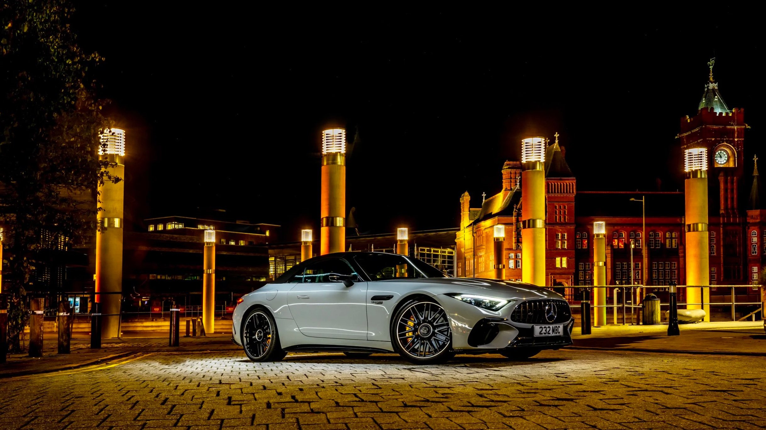 A silver Mercedes-Benz convertible car parked on a cobblestone street at night, illuminated by streetlights with a historic red-brick building and clock tower in the background.