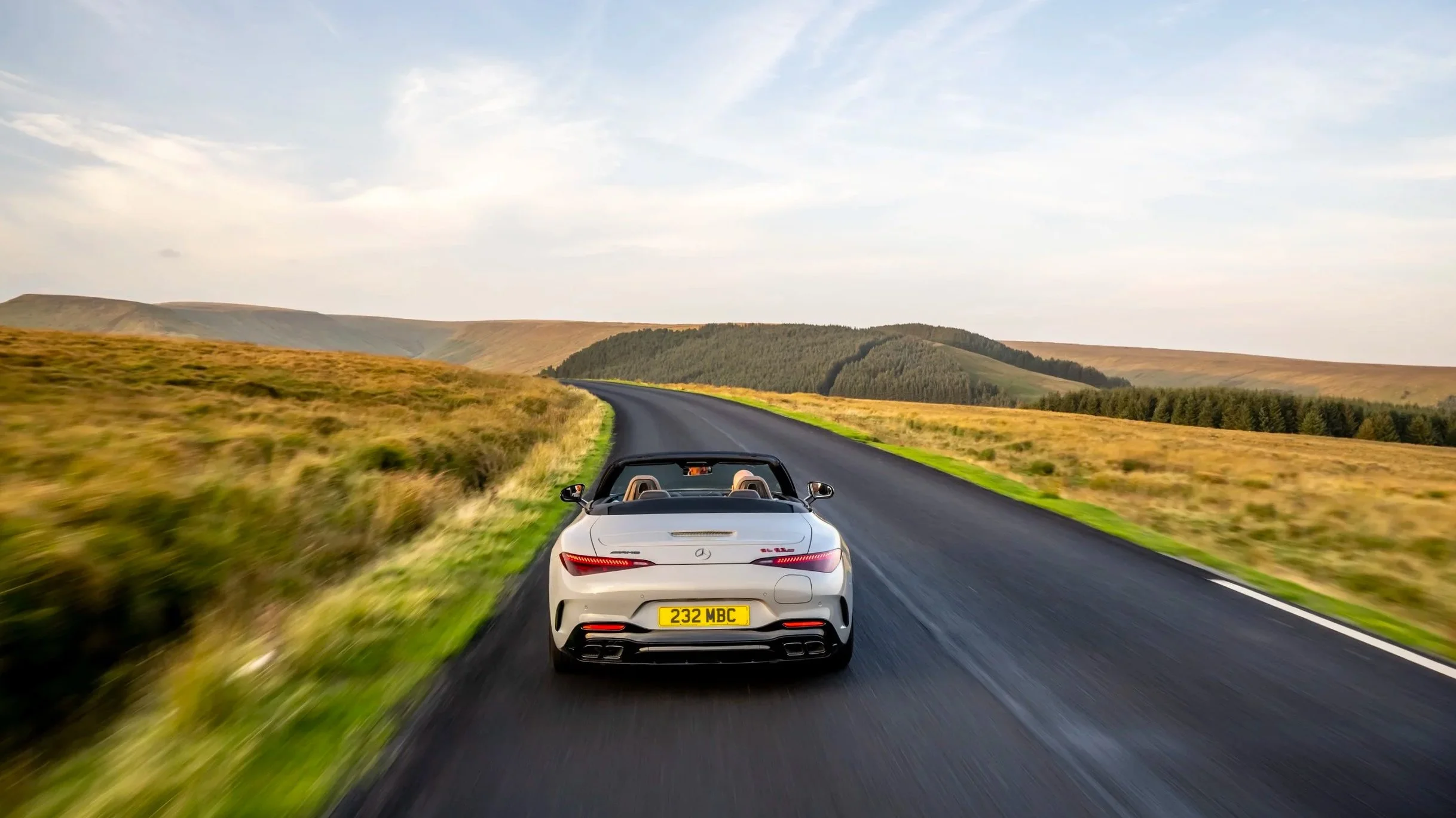 A white convertible sports car driving on a winding rural road through green and yellow fields with hills and trees in the distance under a partly cloudy sky.