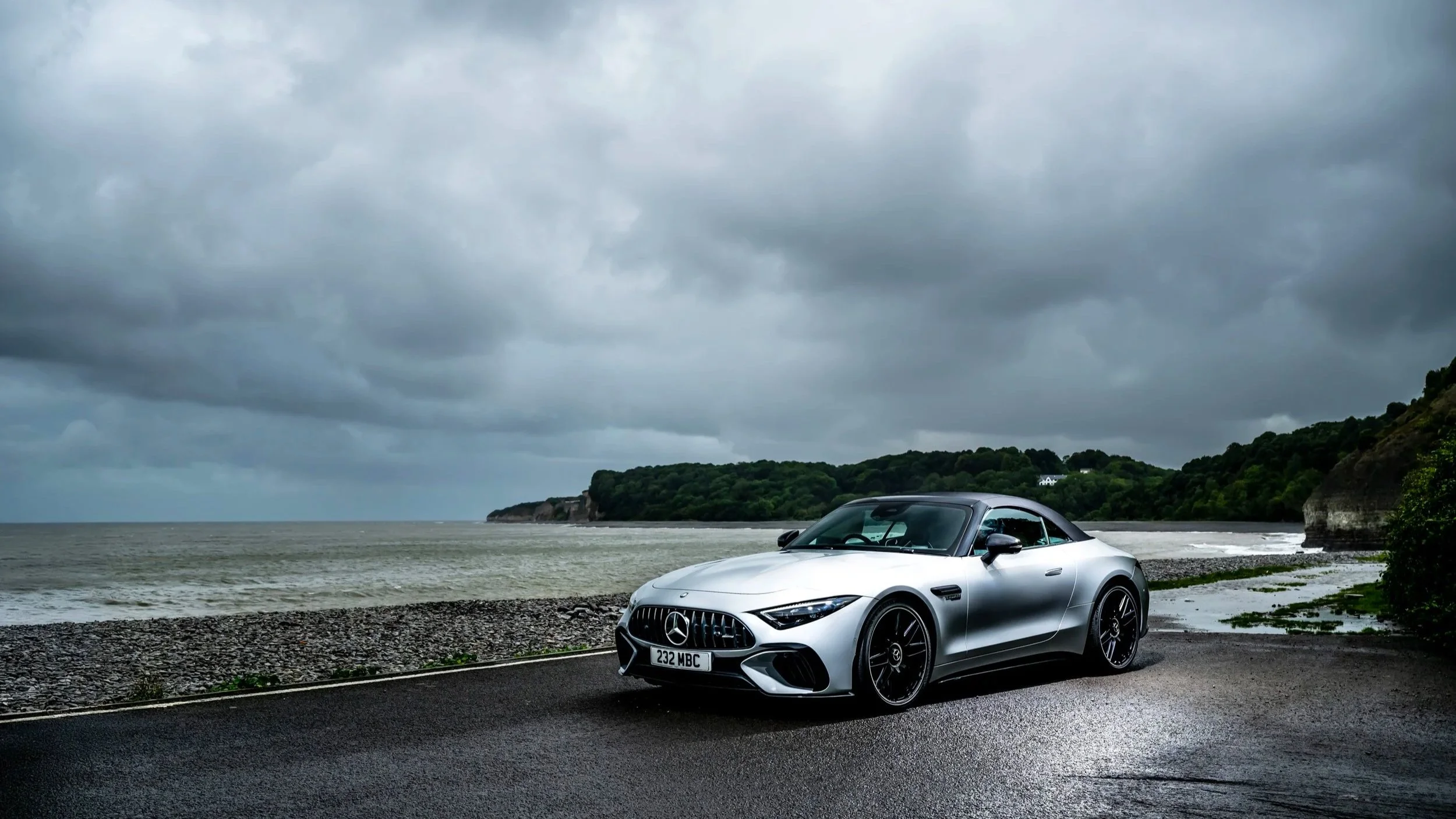 A silver Mercedes-Benz sports car parked along a coastal road with a pebble beach and ocean in the background, under a cloudy sky.