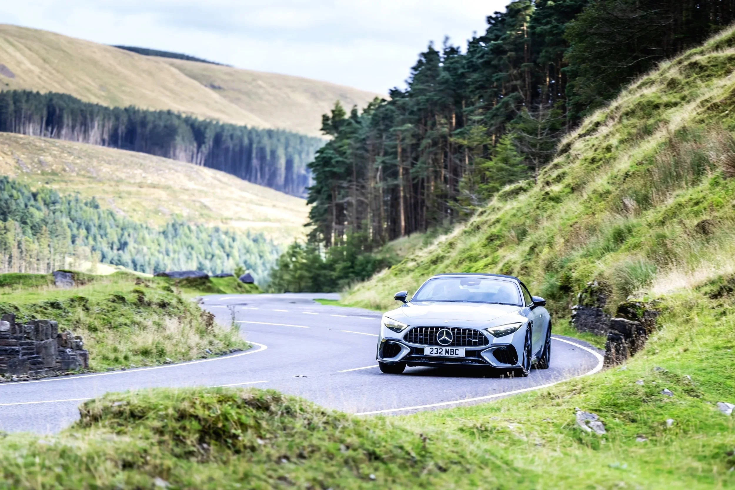 A silver Mercedes-Benz sports car driving on a winding mountain road surrounded by green hills and dense forests.