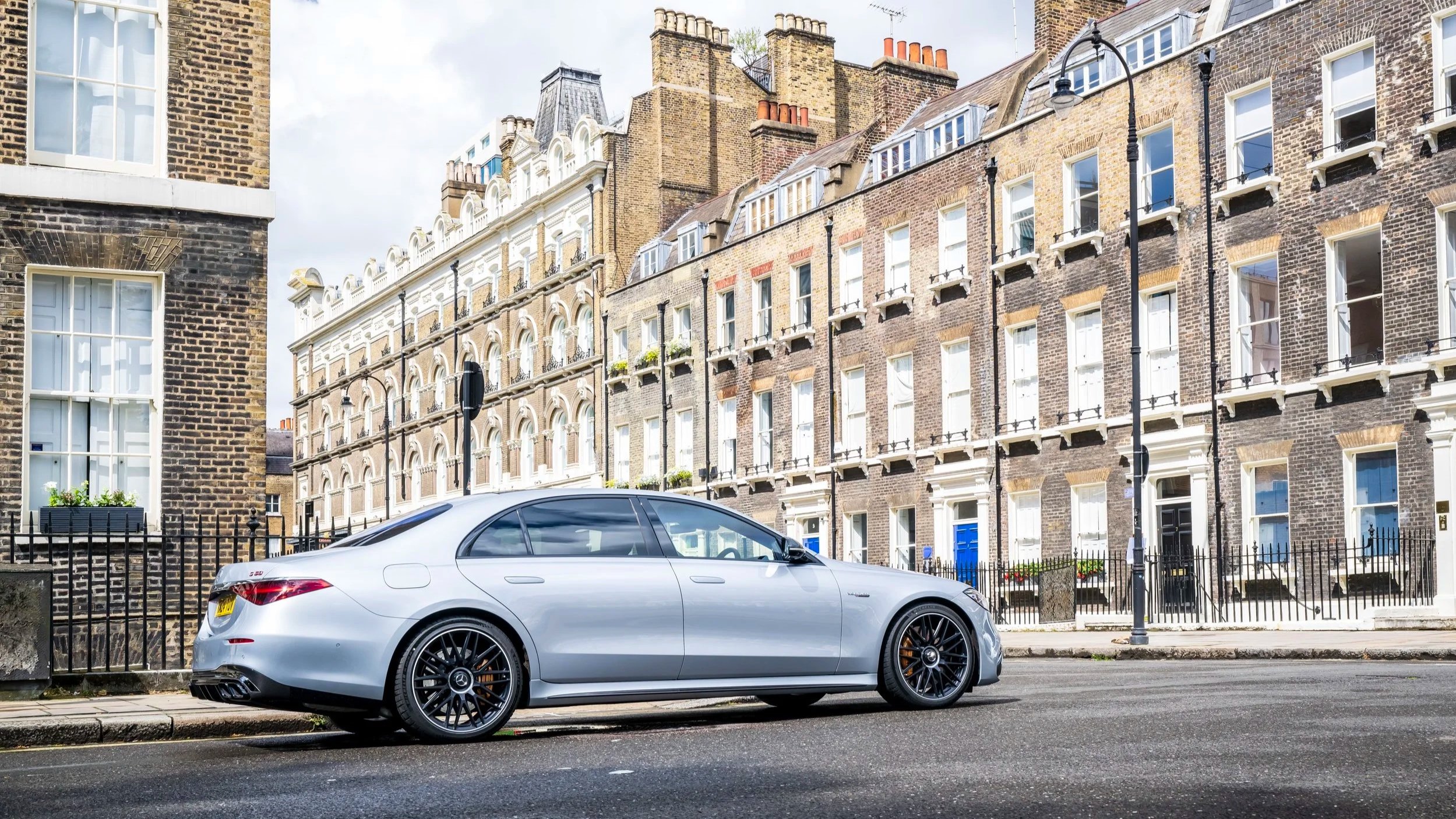 A silver luxury sedan parked on the street in front of classic London-style brick buildings with black iron railings and street lamps.