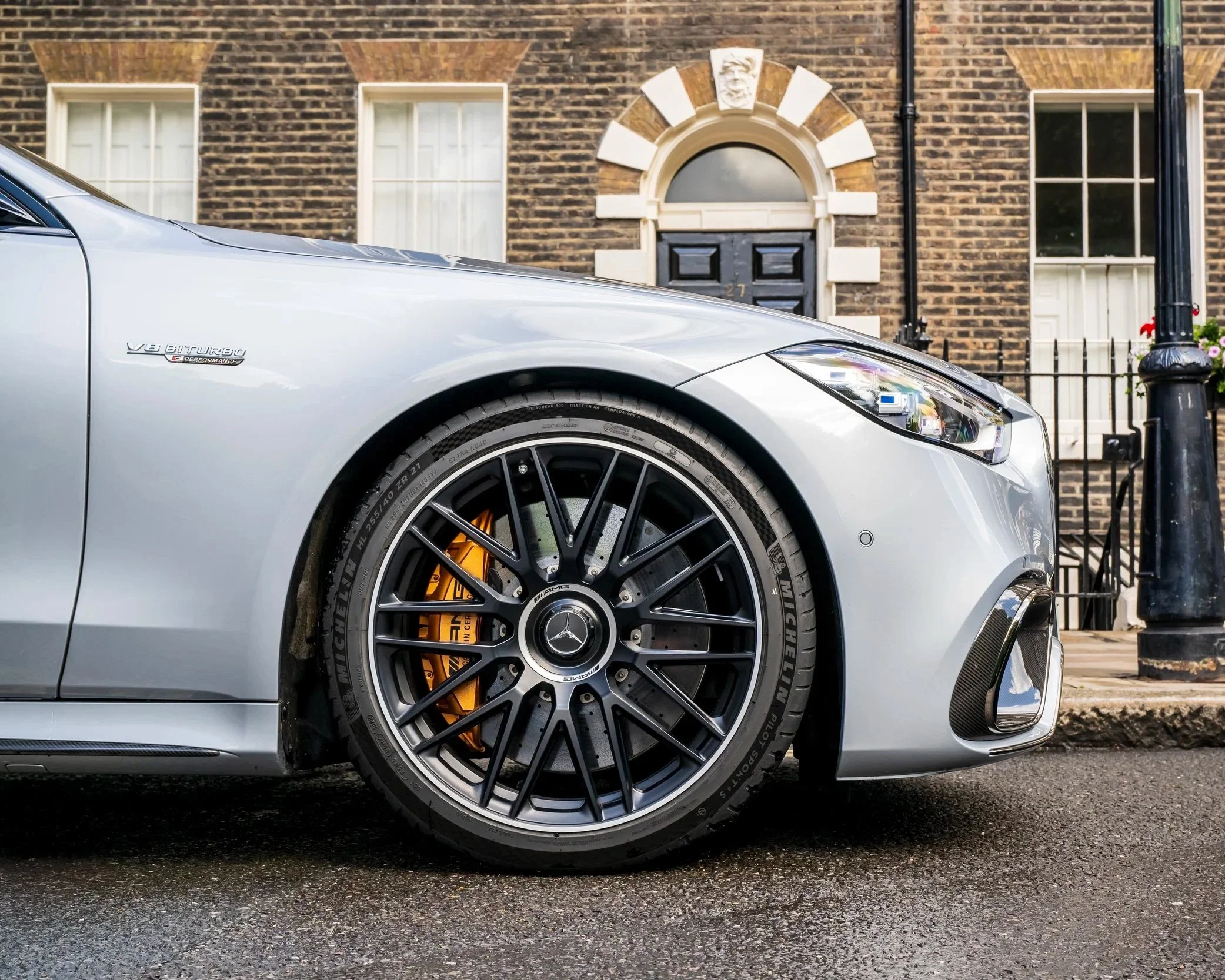 Close-up of a silver Mercedes-Benz luxury car parked on a city street, with a focus on the front wheel and headlight, in front of a brick building with white window frames and a black front door.