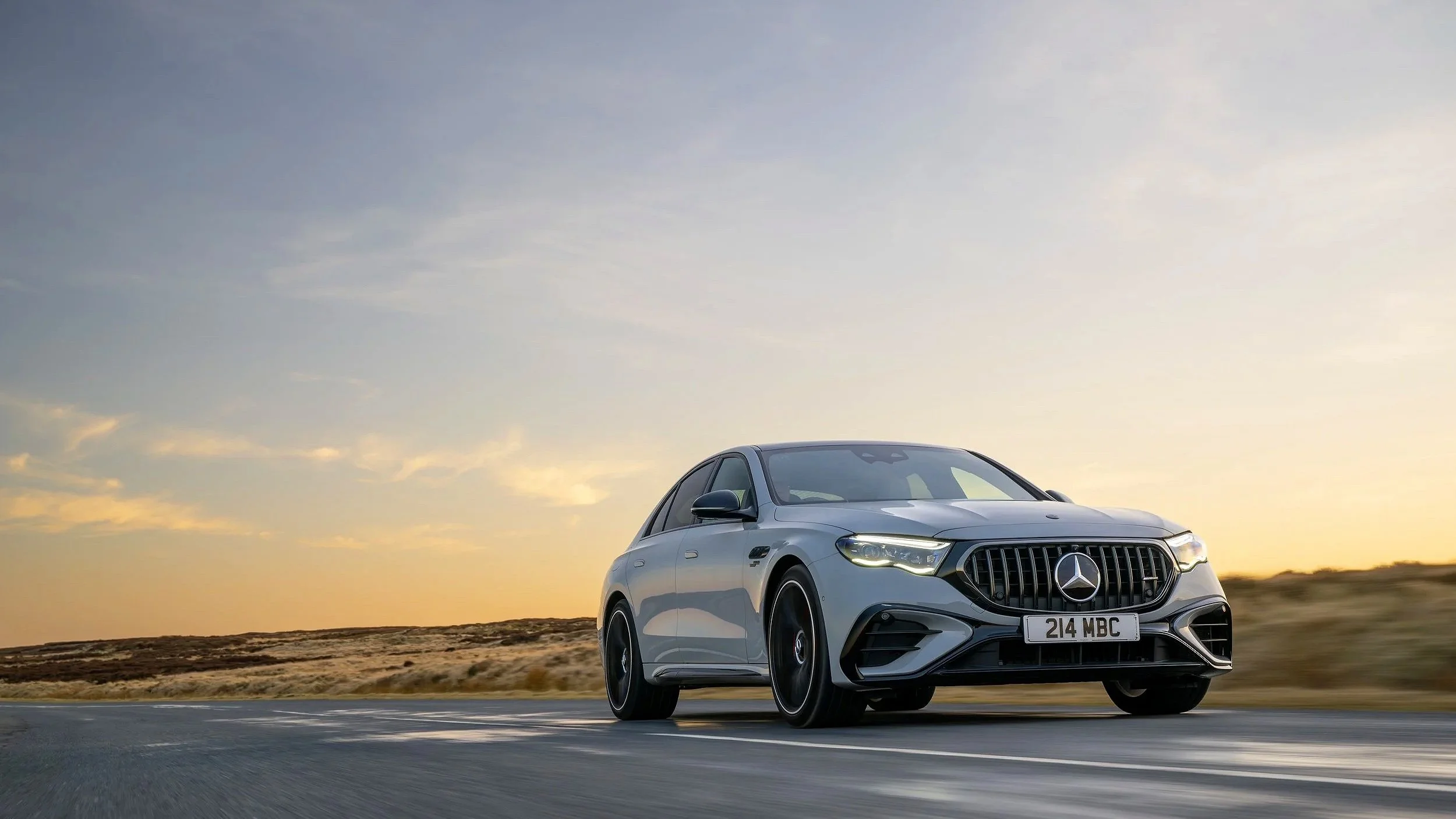 A silver Mercedes-Benz sedan driving on an open road during sunset or sunrise with a partly cloudy sky and dry landscape in the background.