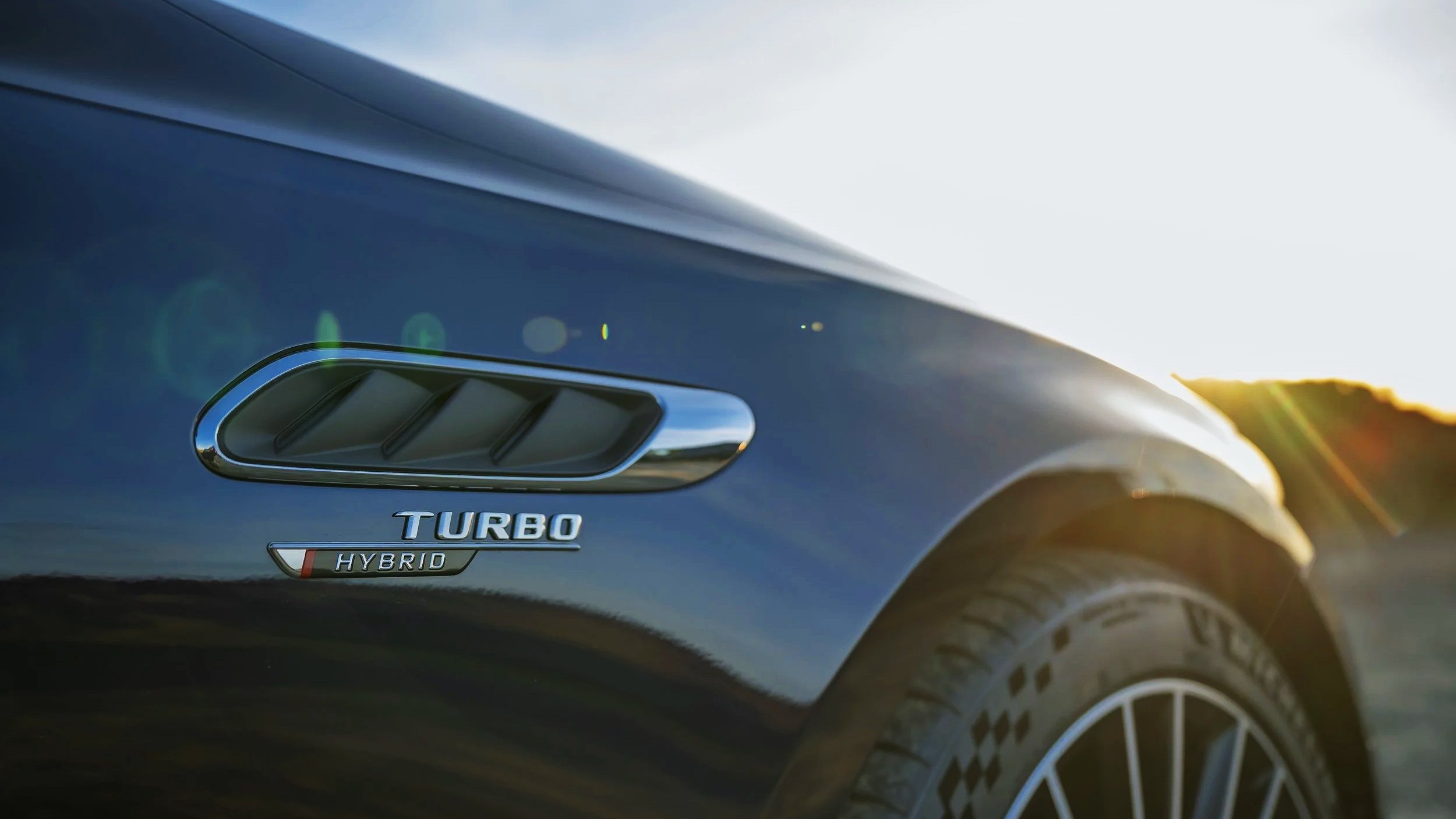 Close-up of a black hybrid turbocharged car with a vent and tire, with sunlight in the background.