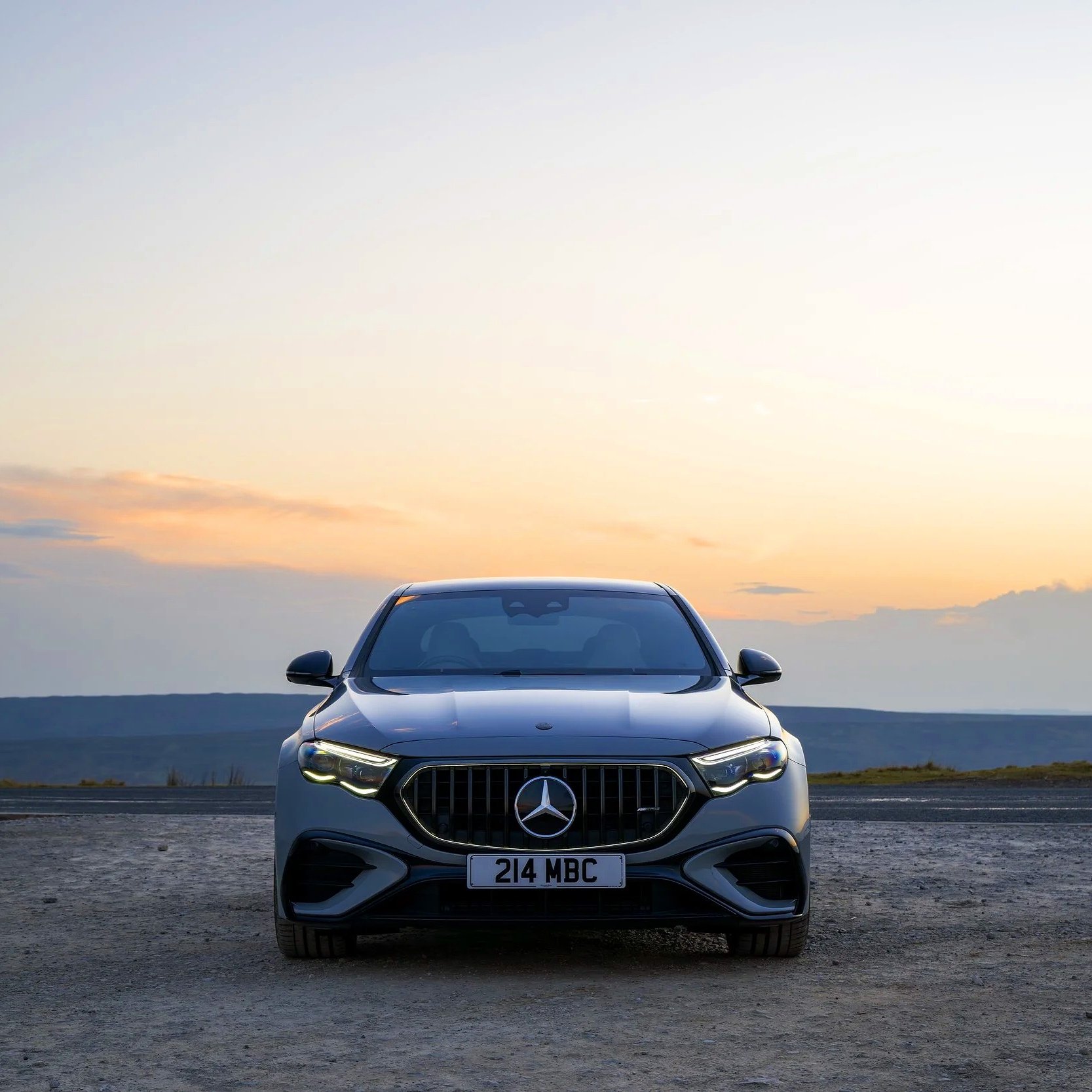 Front view of a black Mercedes-Benz car parked on a gravel surface during sunset, with a scenic background of sky and hills.