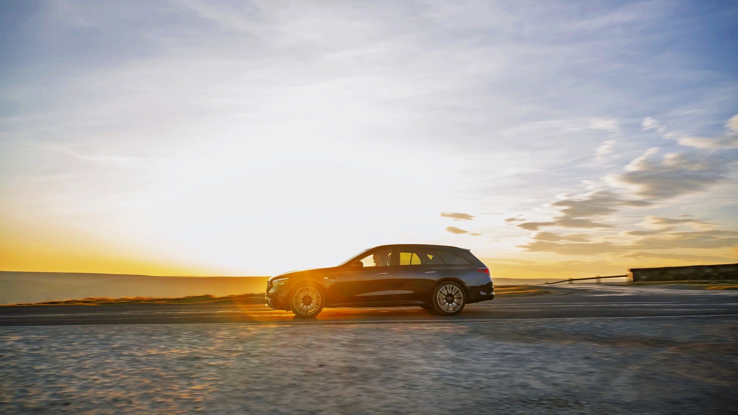 A black station wagon driving on a road during sunset with a partly cloudy sky.