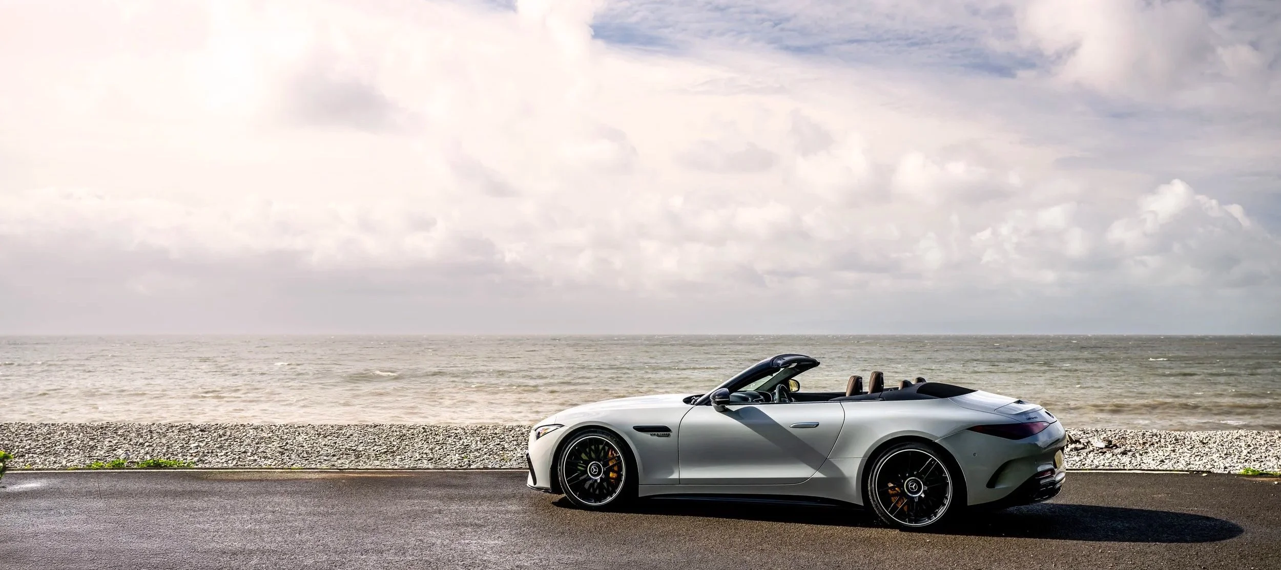 A silver convertible sports car parked on the side of a road near the ocean with a cloudy sky overhead.