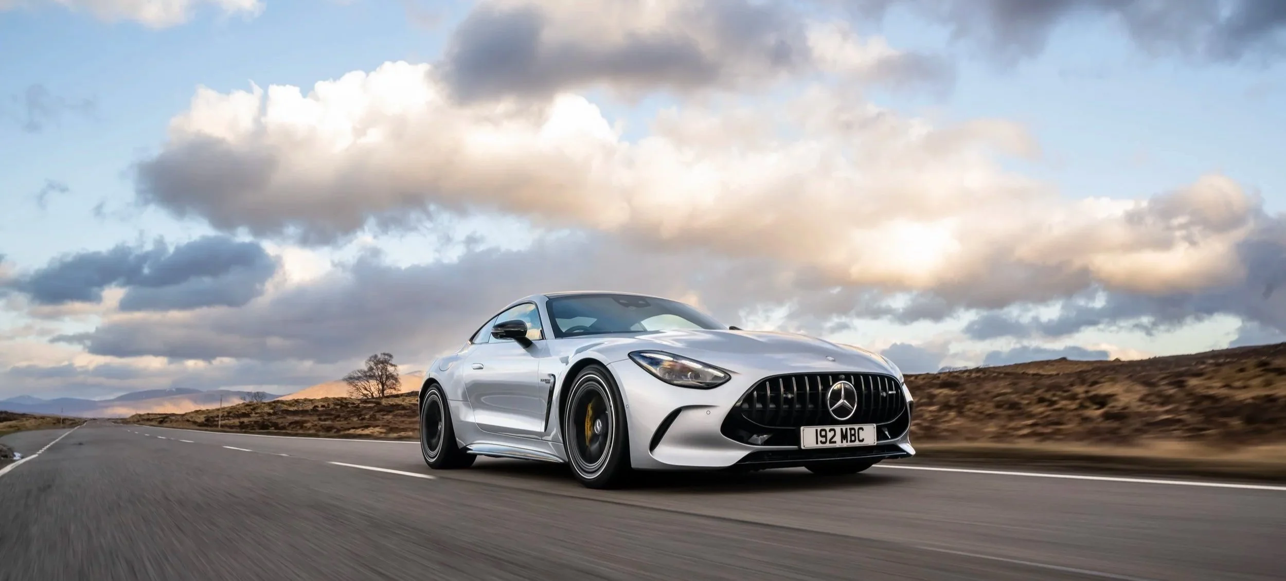 A silver Mercedes sports car driving on a highway through a scenic landscape with mountains, trees, and a partly cloudy sky.