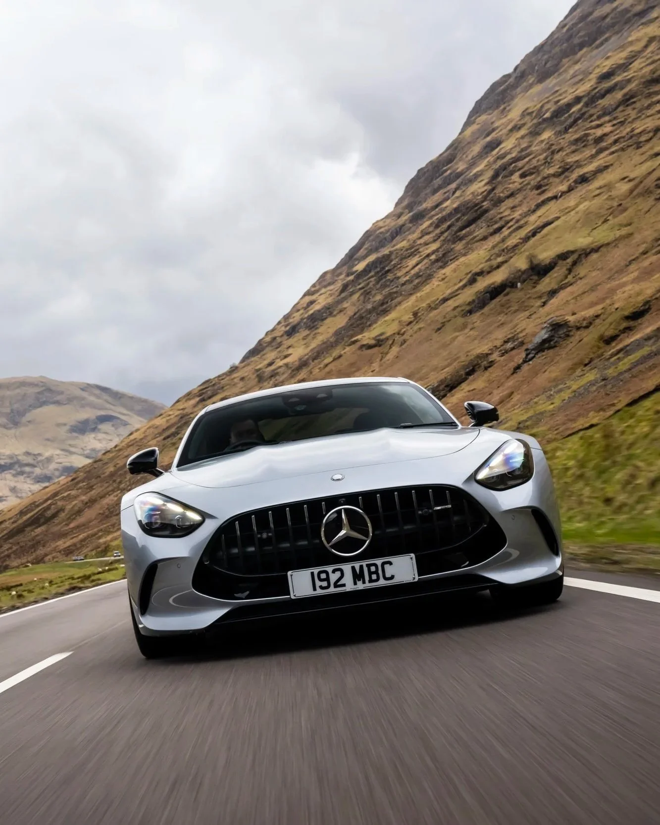 A silver Mercedes-Benz sports car driving on a winding mountain road with grassy hills and rocky slopes, under a cloudy sky.
