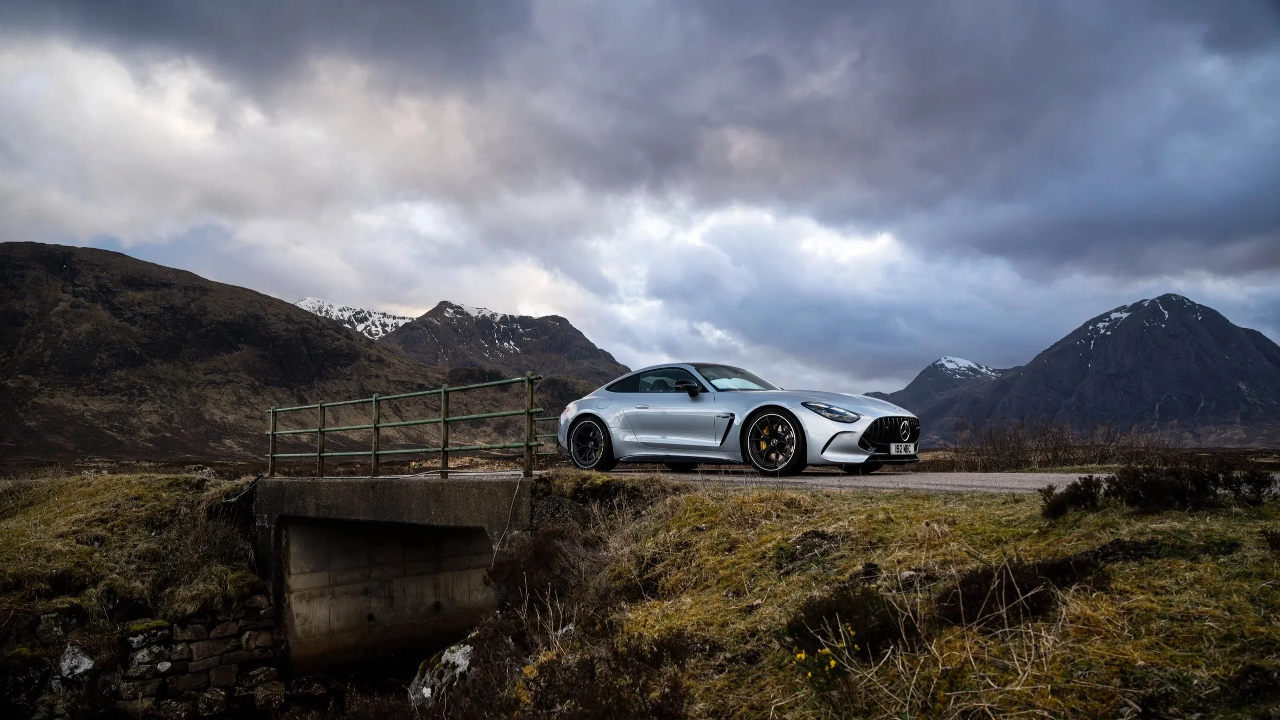 A silver Mercedes-Benz sports car parked on a bridge overlooking a mountainous landscape with snow-capped peaks, under a cloudy sky.