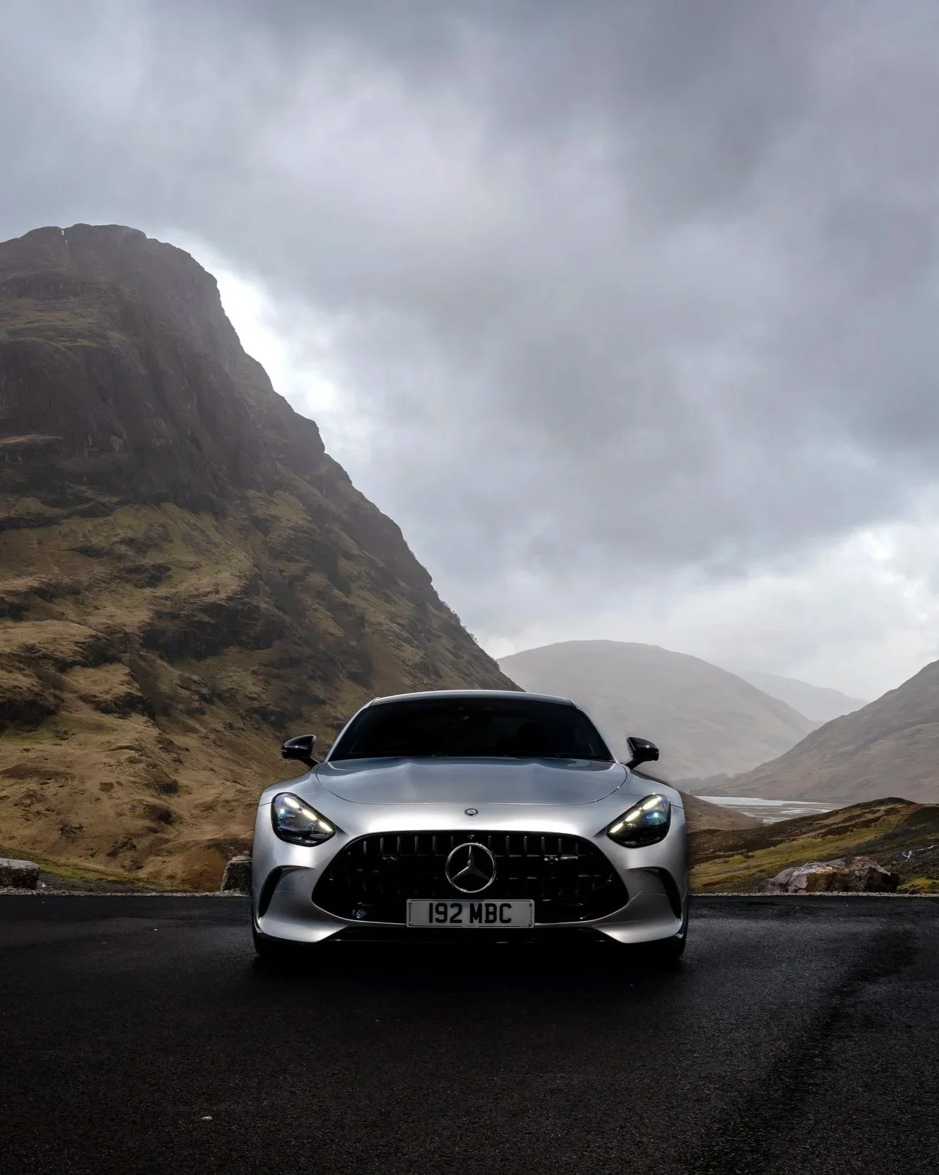 Silver Mercedes-Benz car parked on a dark paved road amidst rugged mountainous landscape with overcast sky.