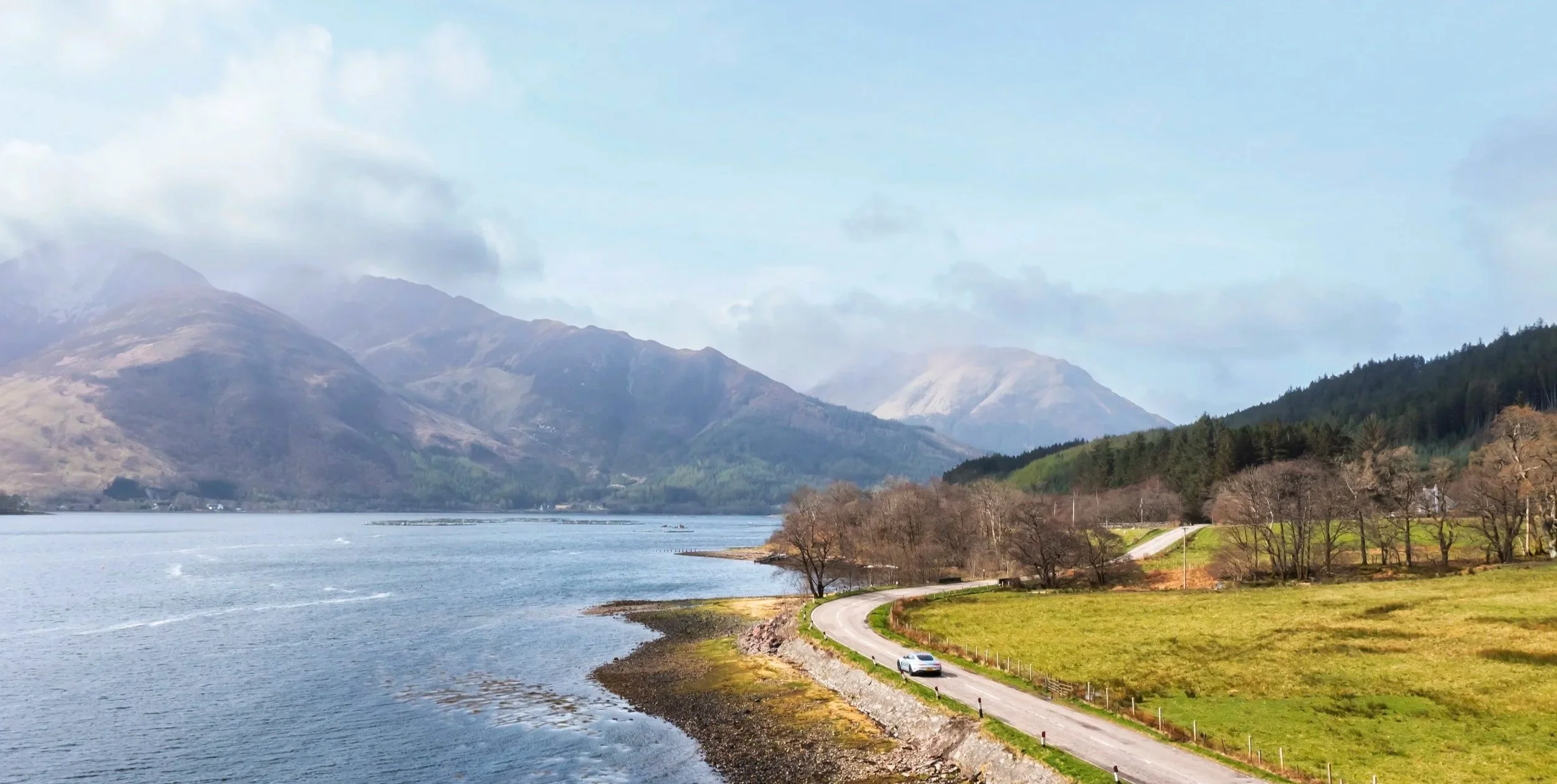 Scenic landscape with a winding road beside a lake, mountains in the background, some trees, and a car traveling on the road.