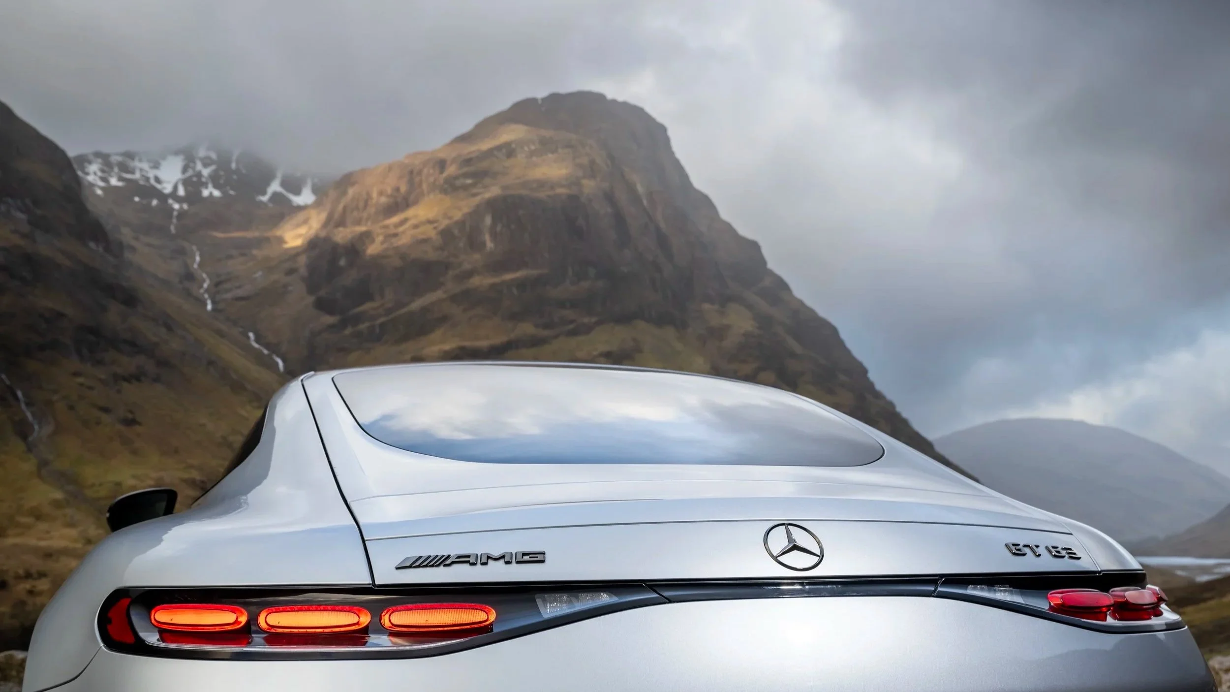 Rear view of a silver Mercedes-Benz AMG GT 63 sports car parked outdoors with mountainous terrain and cloudy sky in the background.