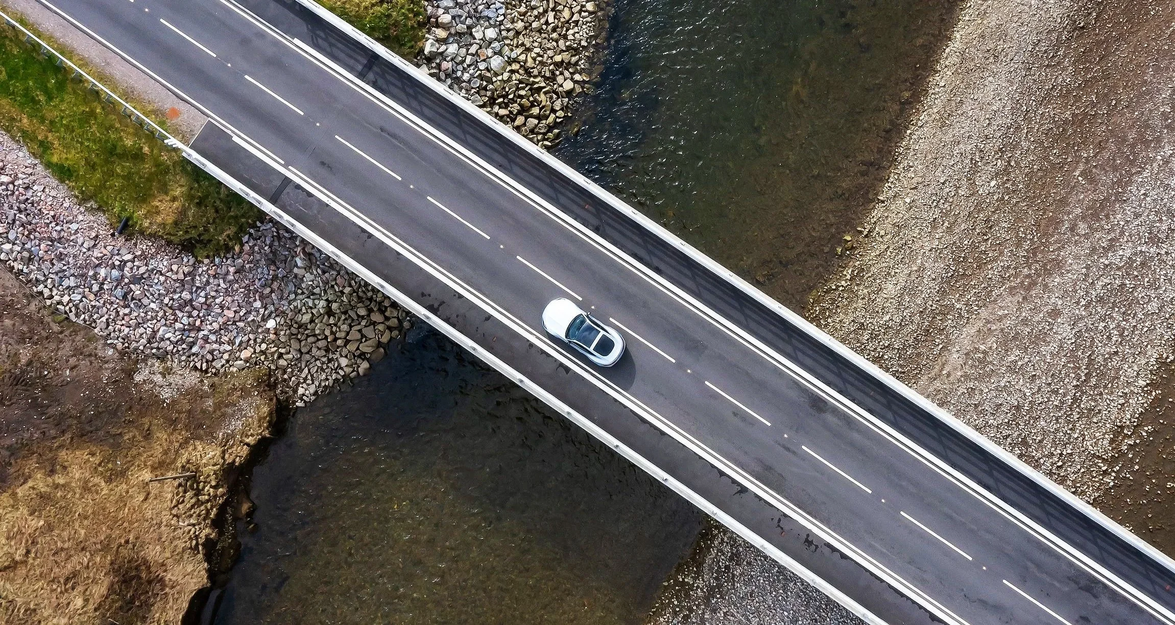 Aerial view of a silver car driving on a bridge over a river, with rocky and grassy banks on either side.