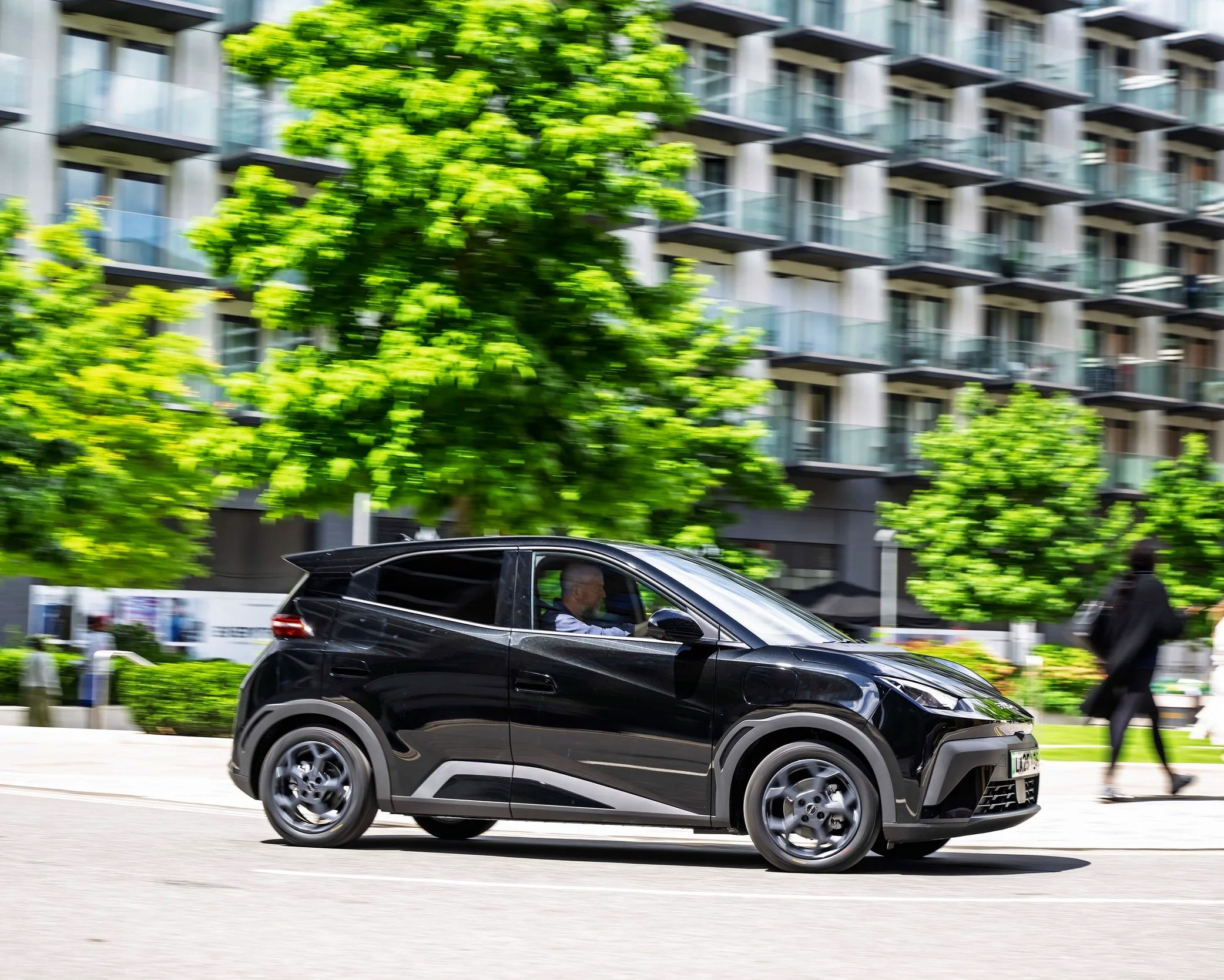 A black compact electric car driving on a city street with a person inside and modern apartment buildings in the background with green trees and pedestrians.