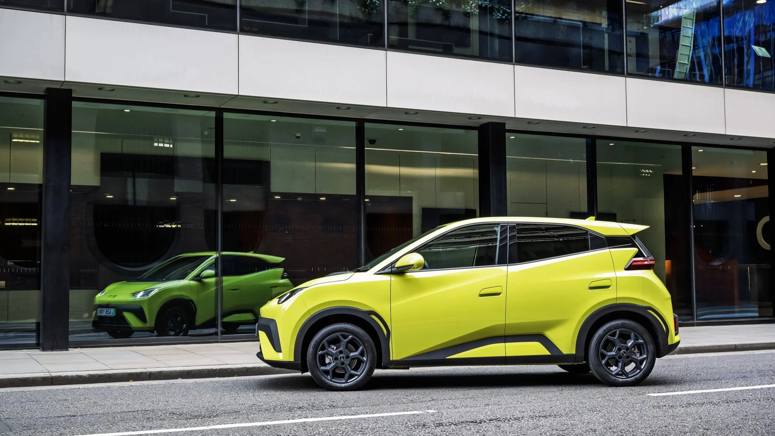 A yellow compact electric SUV parked on the street in front of a modern building with large glass windows, which reflect the vehicle and part of another green car inside.