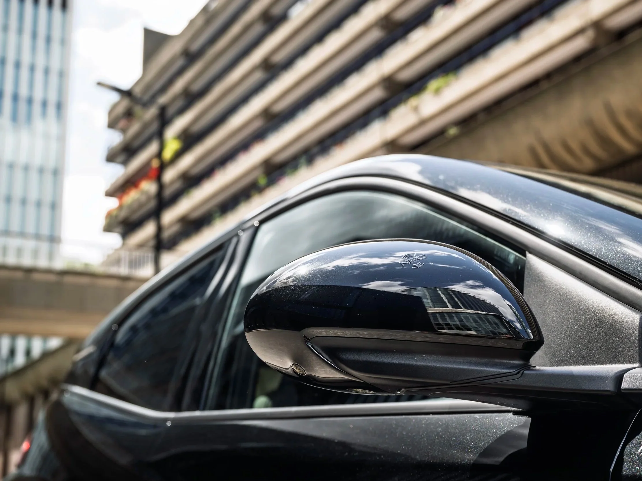 Close-up of a black car’s side mirror with a building, balcony, and sky reflected in the mirror, and a blurred urban background.
