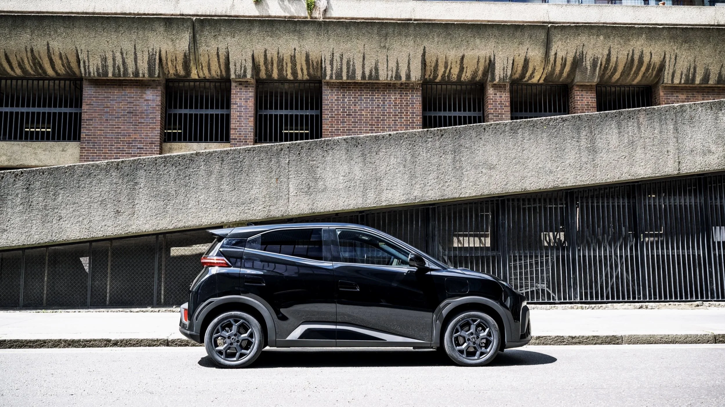 A black compact electric car parked on the street in front of a building with brick and concrete walls and barred windows.