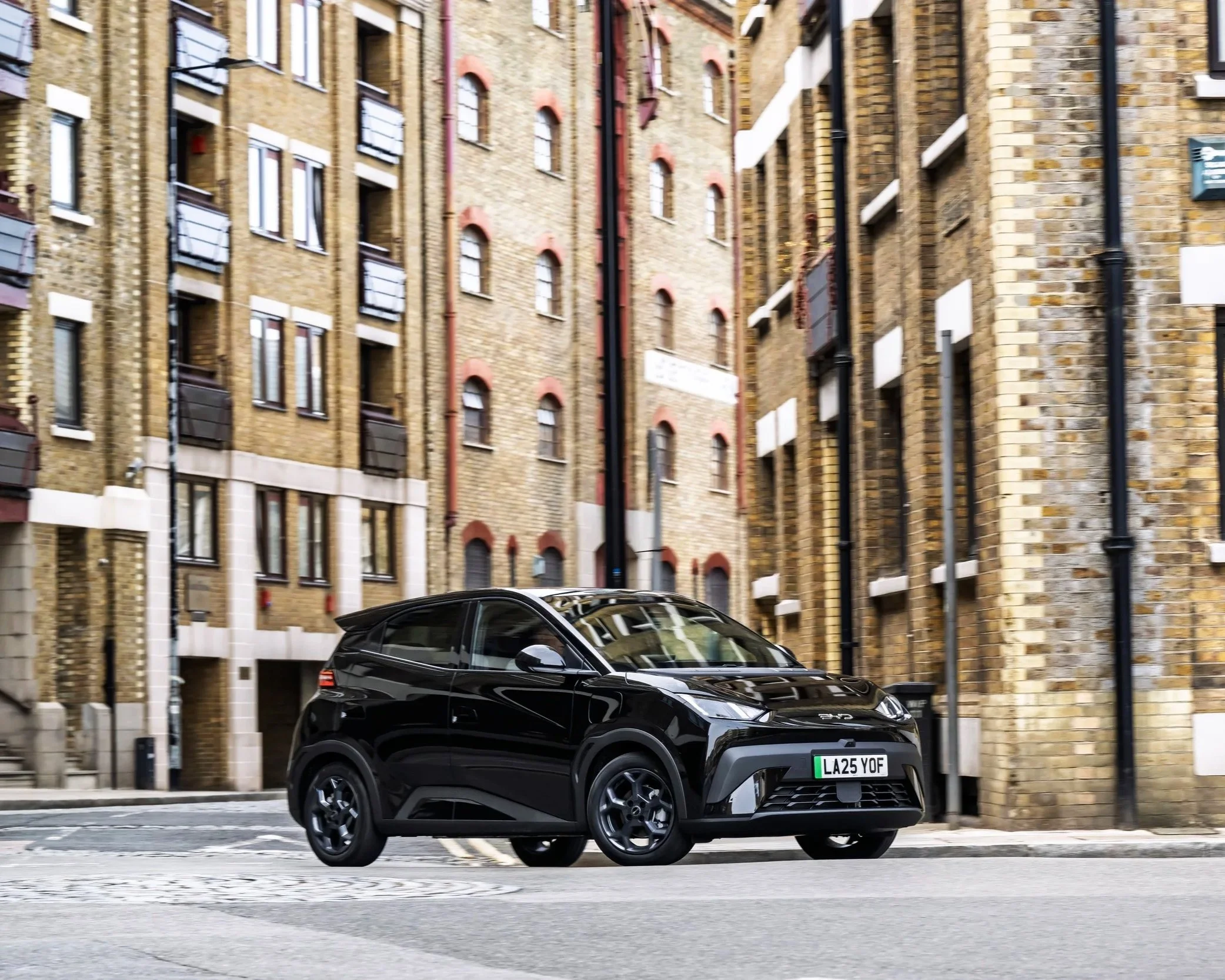 A black electric car parked on a city street with tall brick apartment buildings in the background.