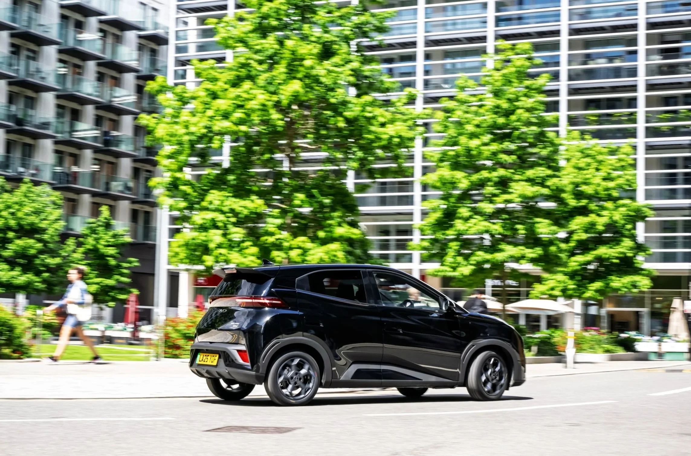 A black compact SUV driving on a city street with buildings, green trees, and pedestrians in the background.