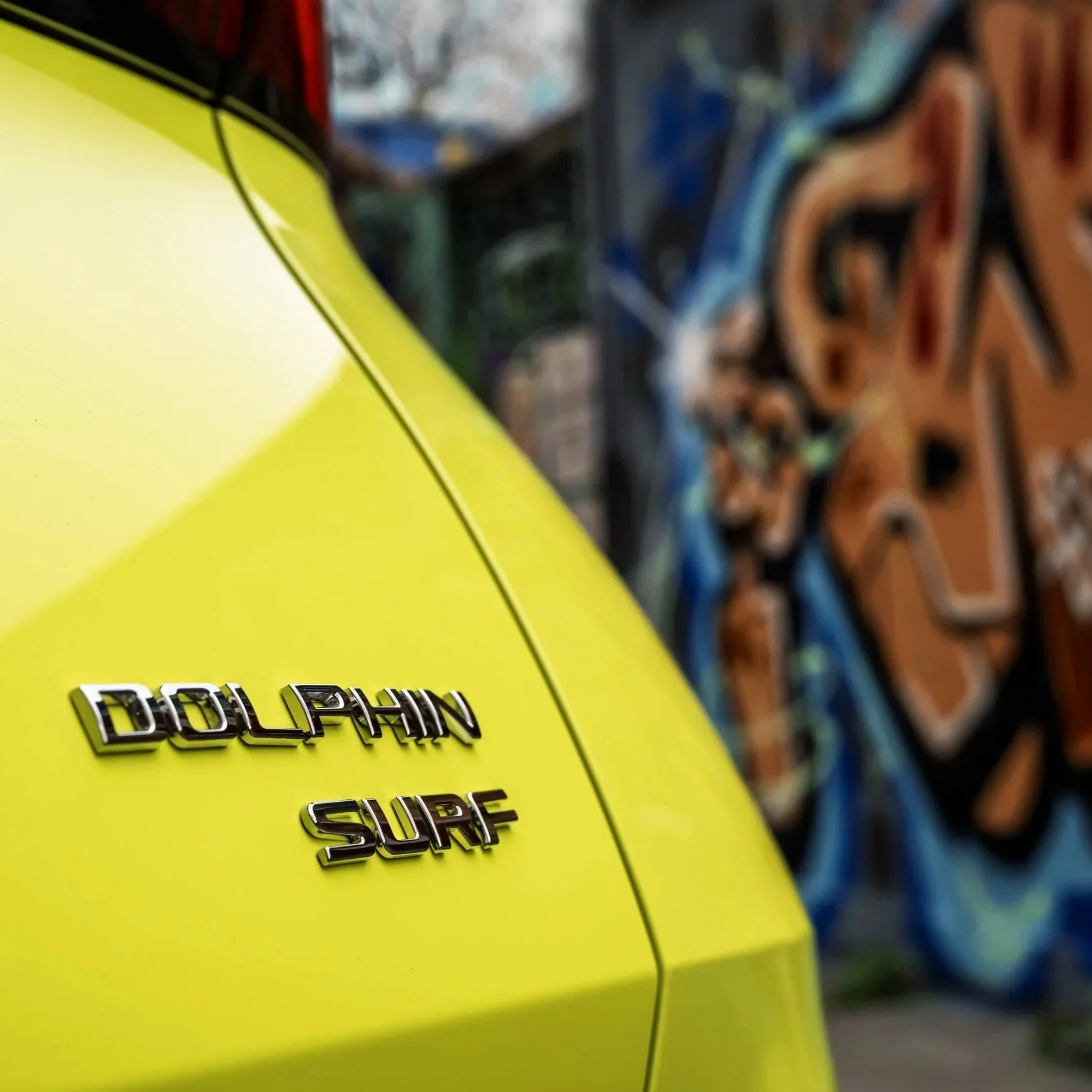 Close-up of a yellow vehicle with chrome emblems that read `DOLPHIN` and `SURF` on its side, with graffiti art in the background.