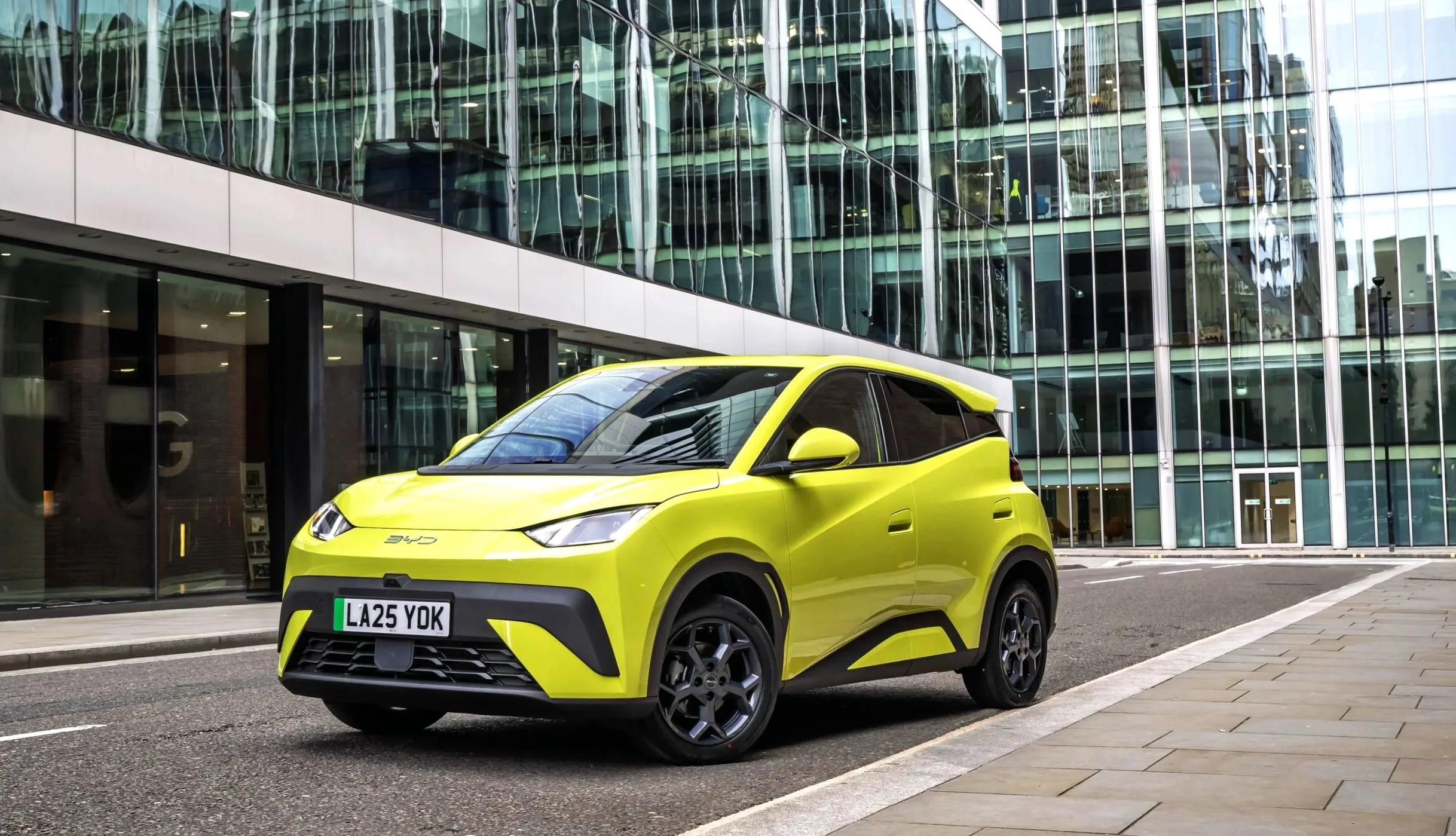 A bright yellow compact electric car parked on an urban street in front of a modern glass office building.