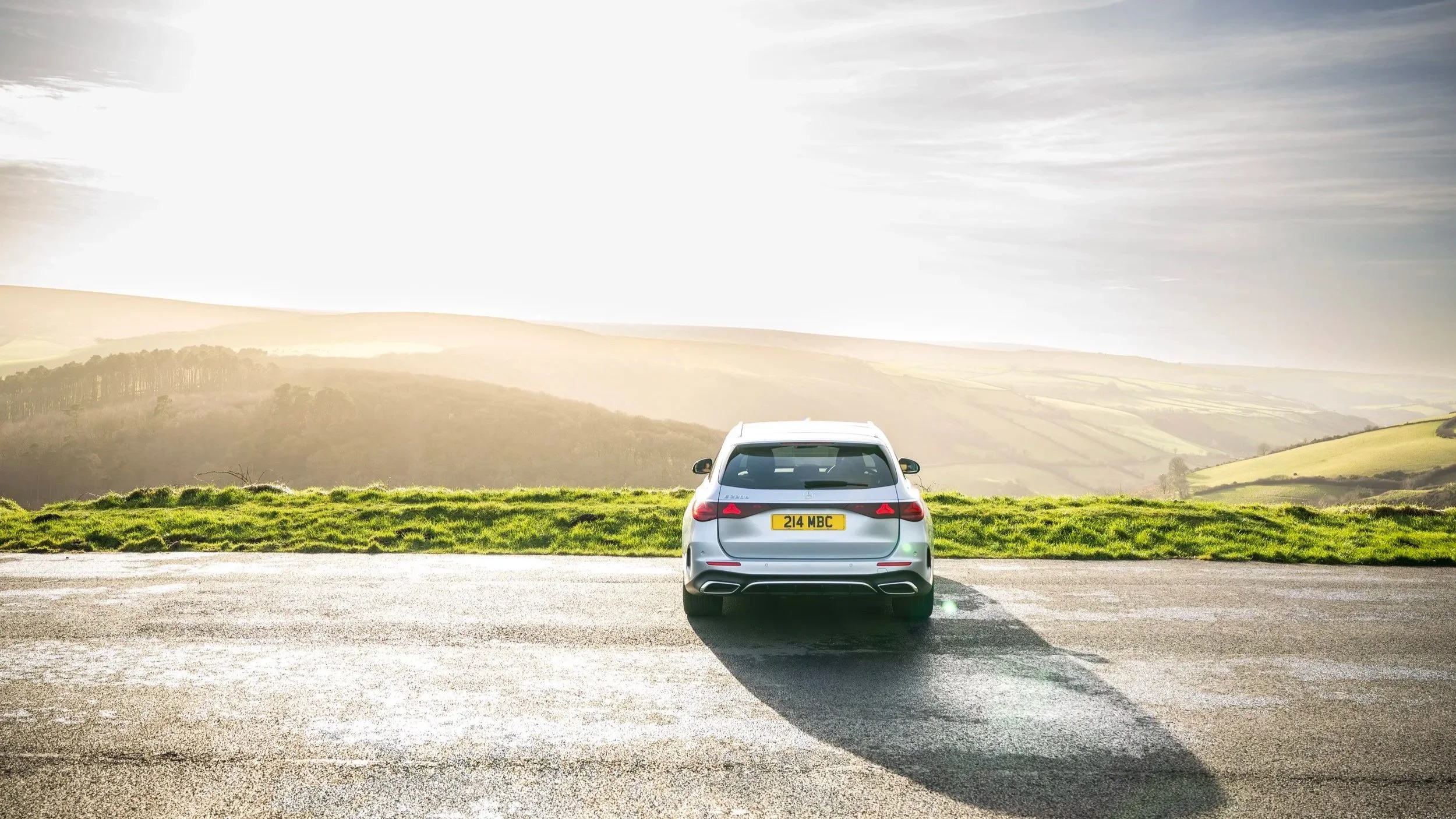 A silver car parked on an empty roadside overlooking a distant landscape of rolling hills and a partly cloudy sky, with the sun shining brightly.