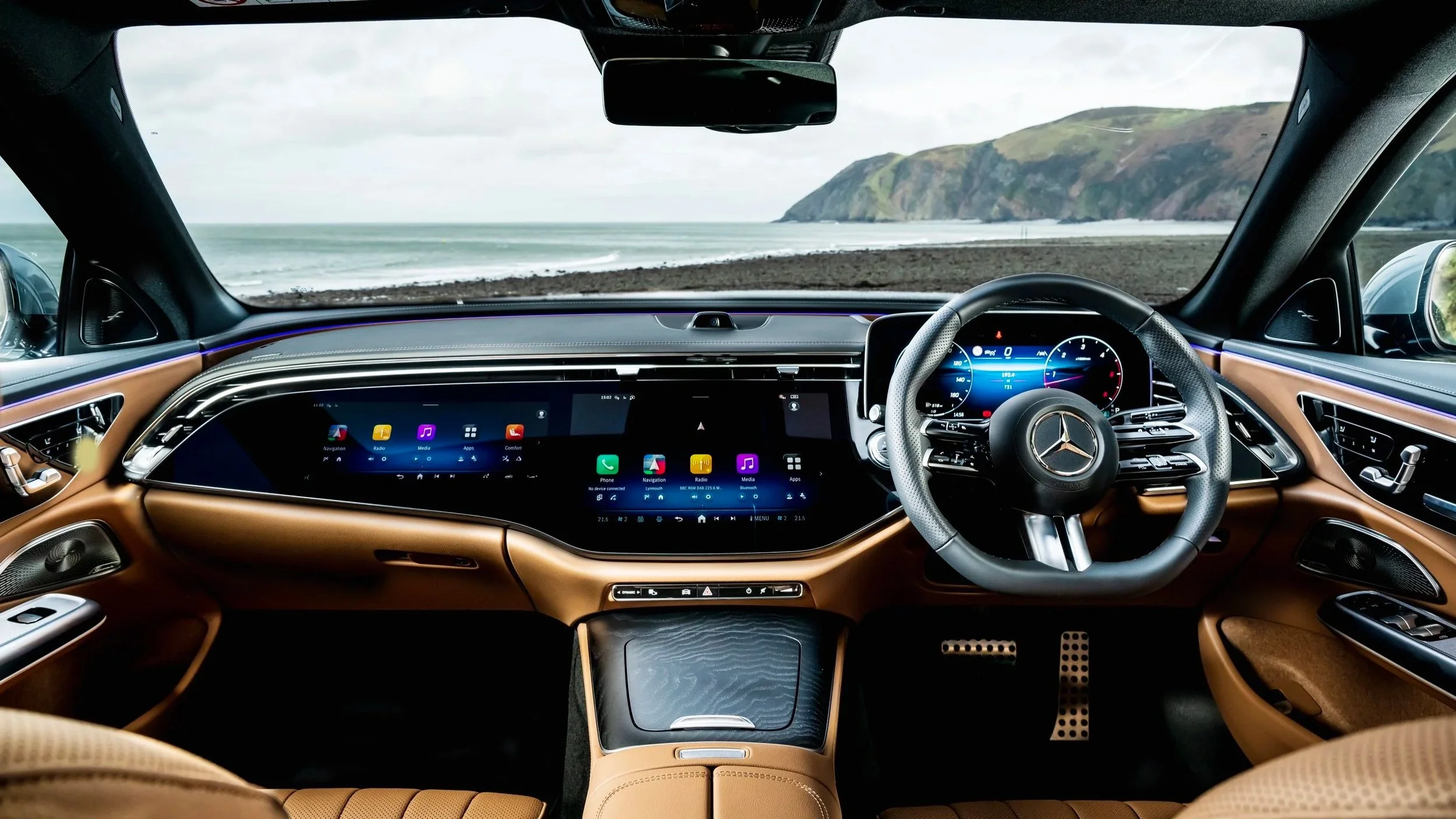 Inside the cabin of a Mercedes-Benz vehicle, showing a sleek digital dashboard, leather-wrapped steering wheel with Mercedes logo, and a panoramic view of a coastline with a beach and green hills through the windshield.