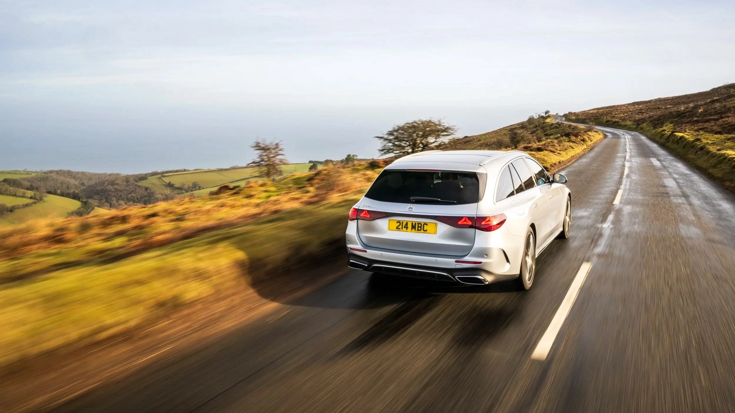 A white station wagon driving on a wet, winding country road through rolling hills with sparse trees and golden grass, under a cloudy sky.