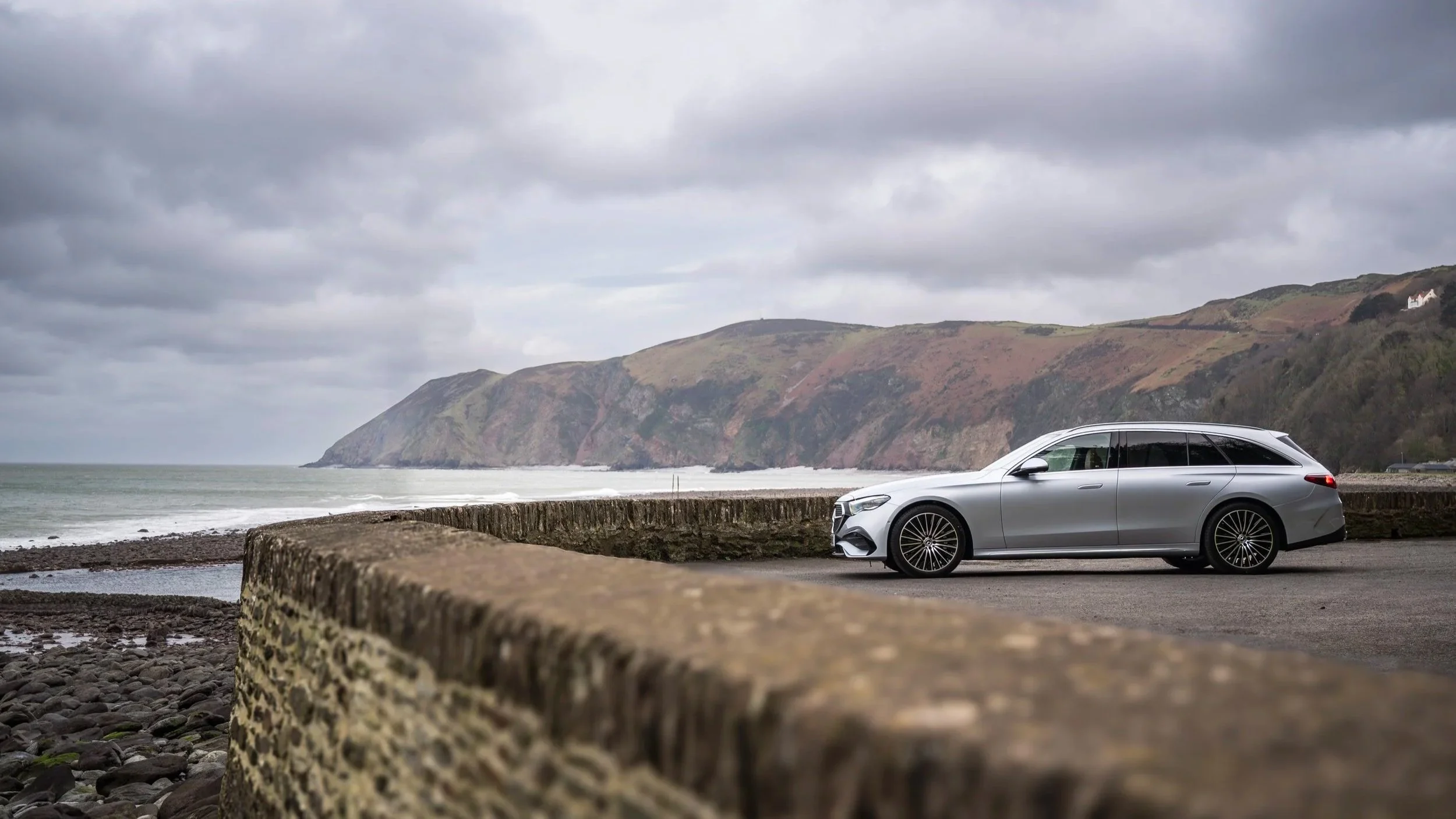 A silver station wagon parked on a coastal road with rocky shoreline, cliffs in the background, and a cloudy sky.