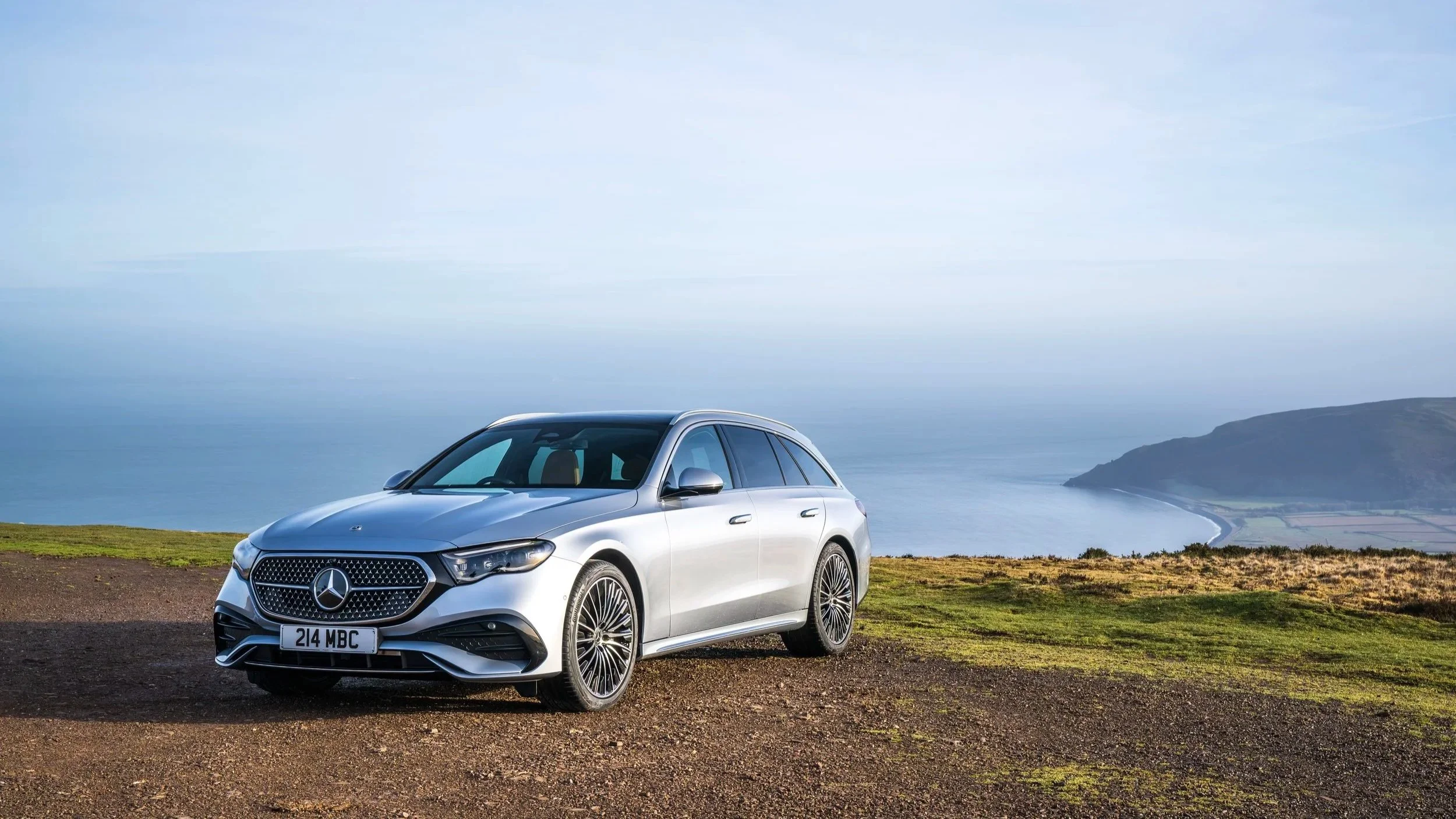 A silver Mercedes-Benz station wagon parked on a hilltop overlooking a coastal landscape with the ocean and cliffs in the background.