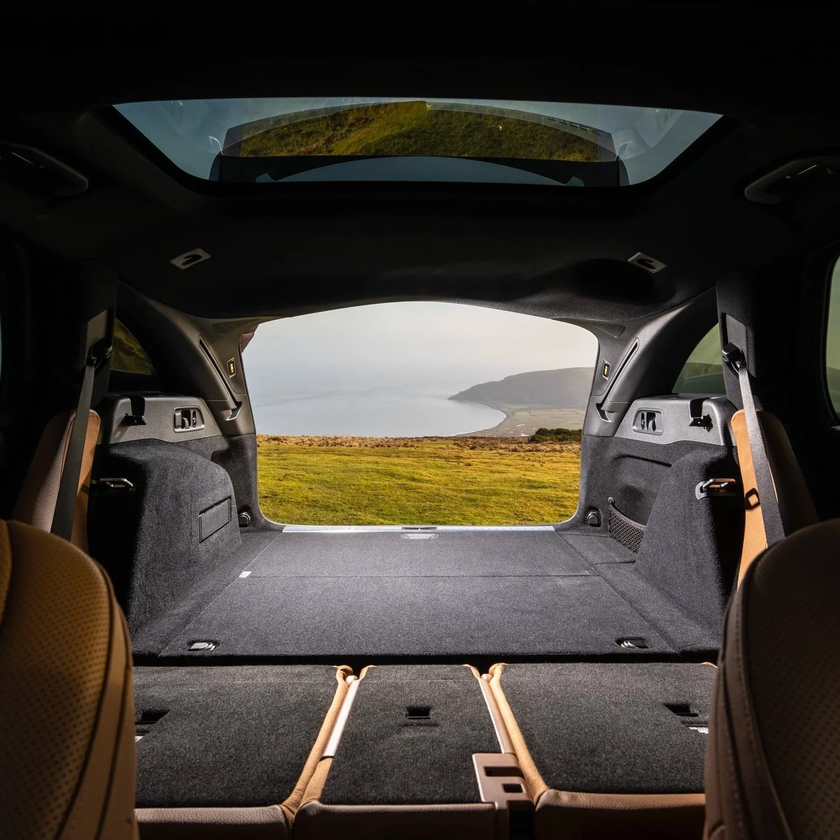 View through the open rear of an SUV showing a scenic landscape with grassy fields, water, and hills or mountains in the distance.