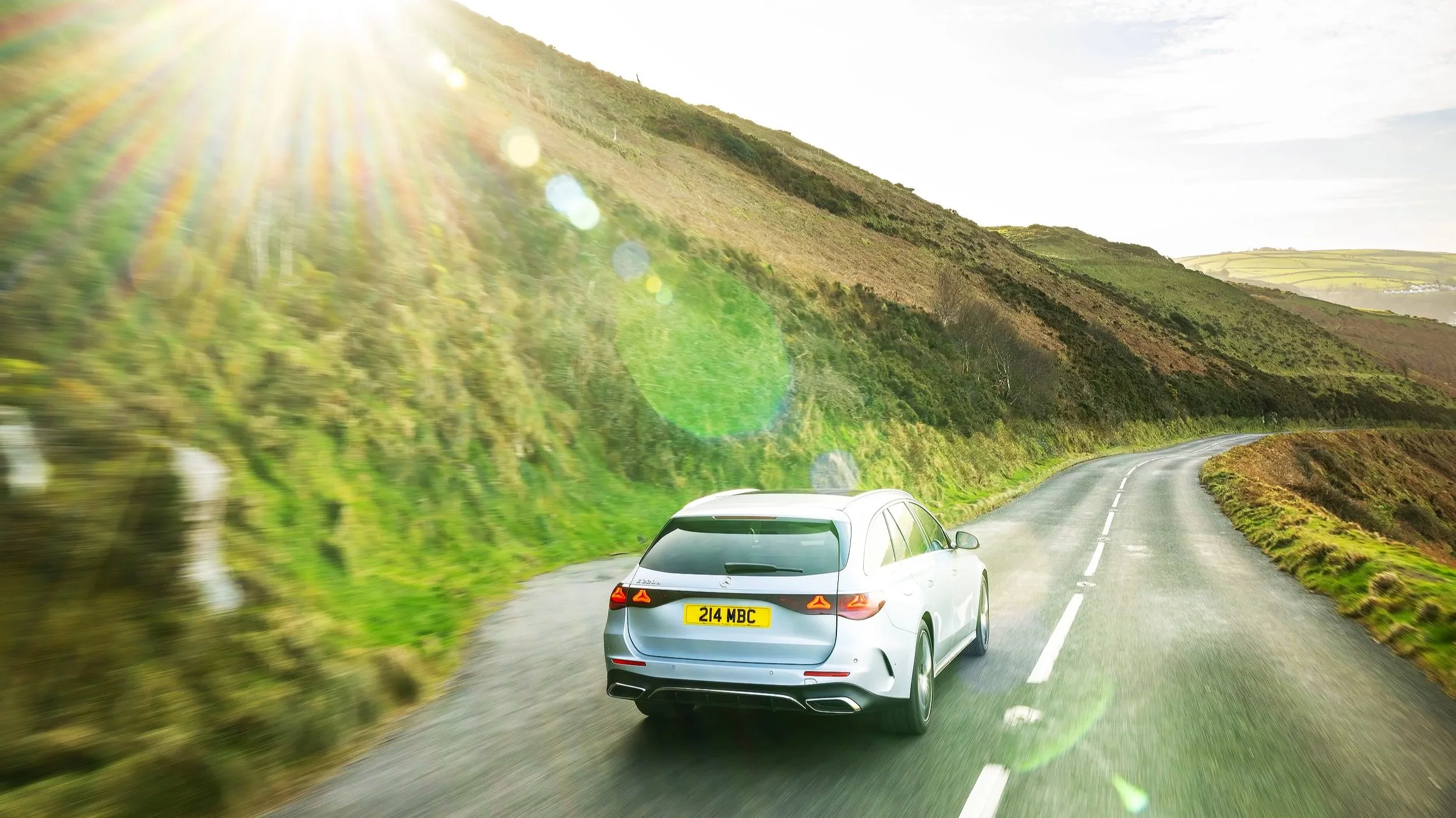 A silver car driving on a winding road through green, rolling hills during daylight with sunlight glare and lens flare effects.
