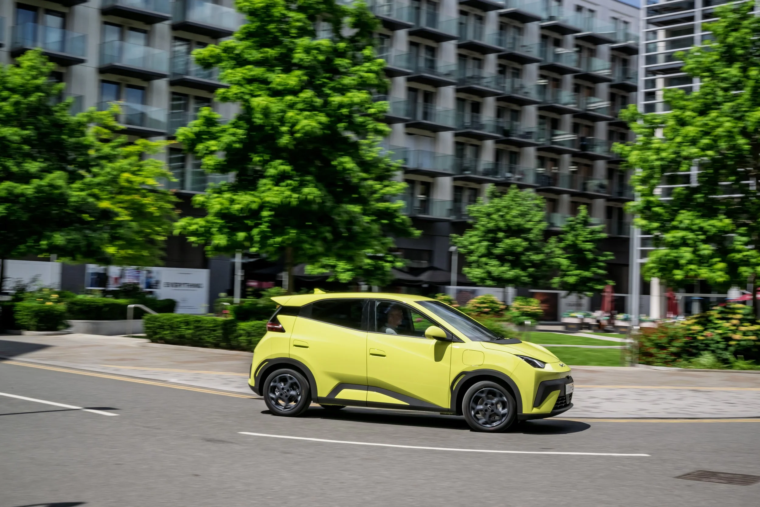 A yellow compact electric car driving on a city street with apartment buildings and green trees in the background.