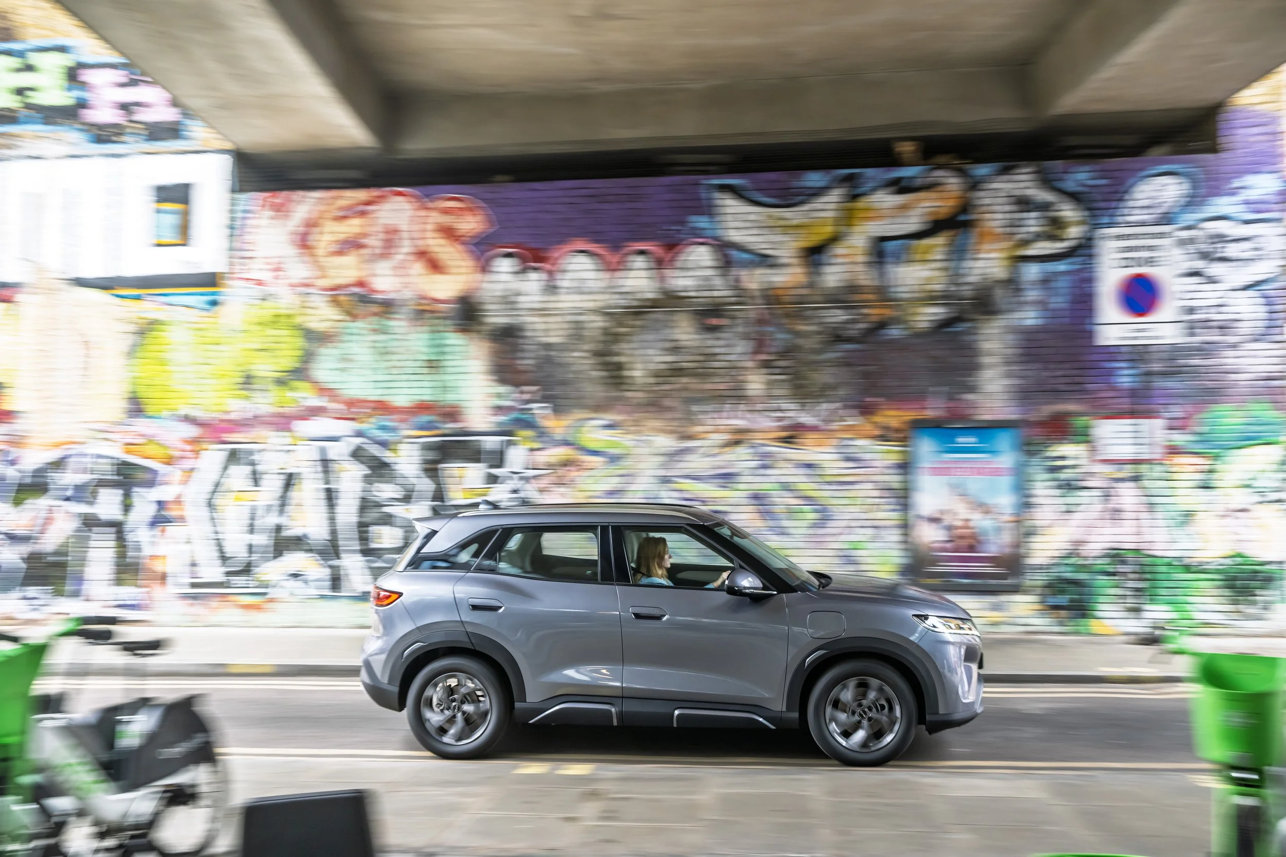 Moving gray SUV driving on city street with vibrant graffiti-covered wall in background.