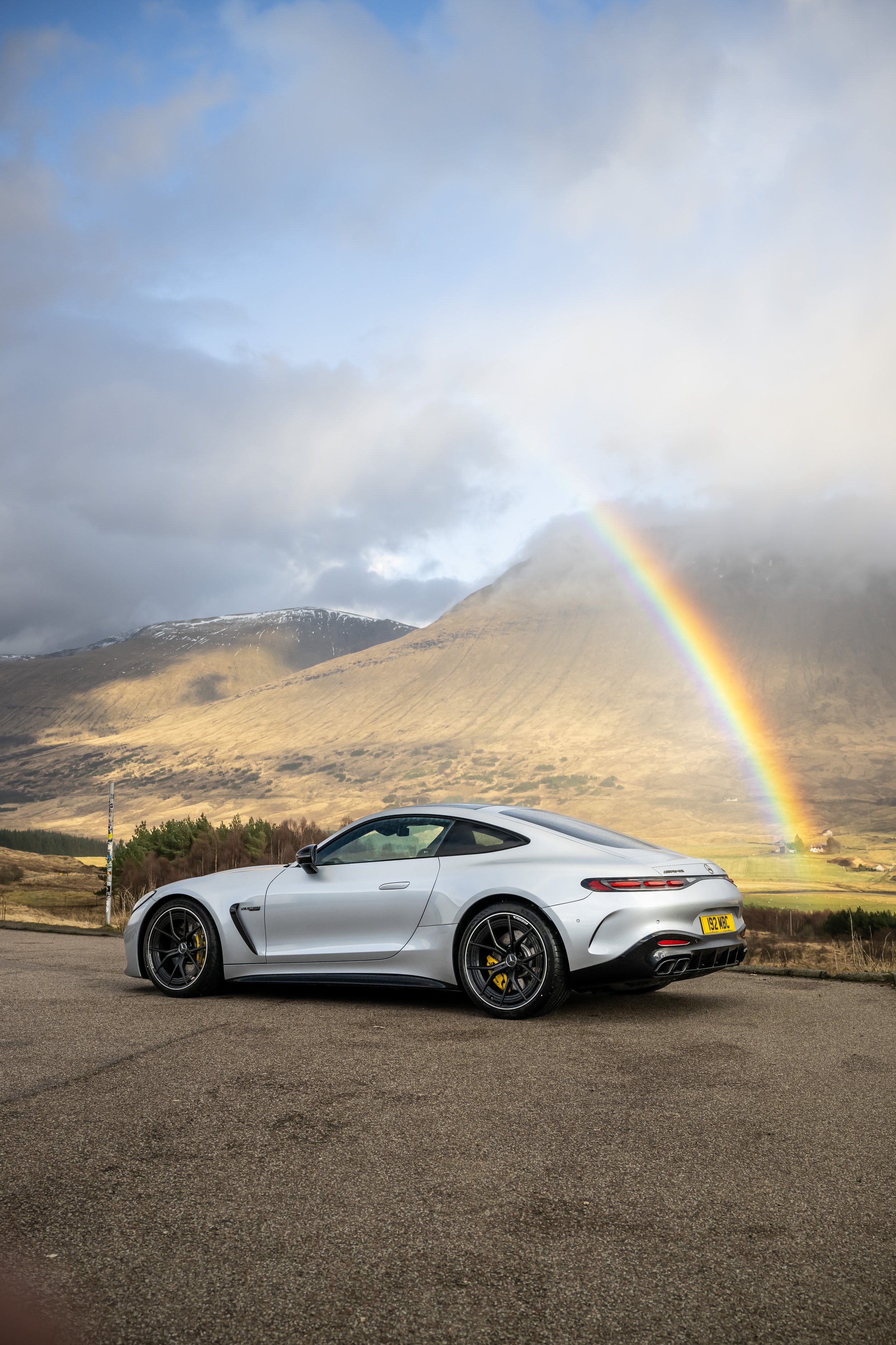 Silver sports car parked on a paved road with a mountain and rainbow in the background